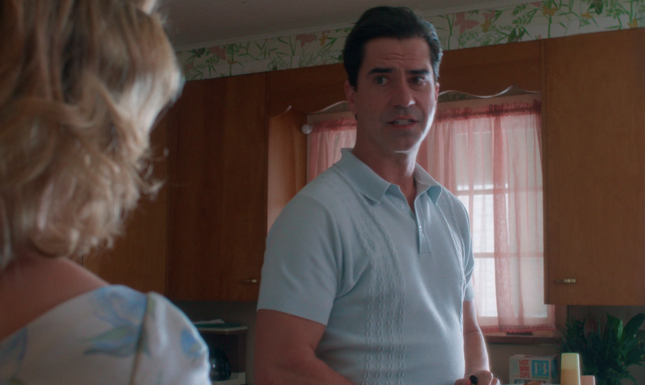A dark-haired man in a light blue polo shirt stands in a vintage kitchen, looking intently at a woman whose back is to the camera.