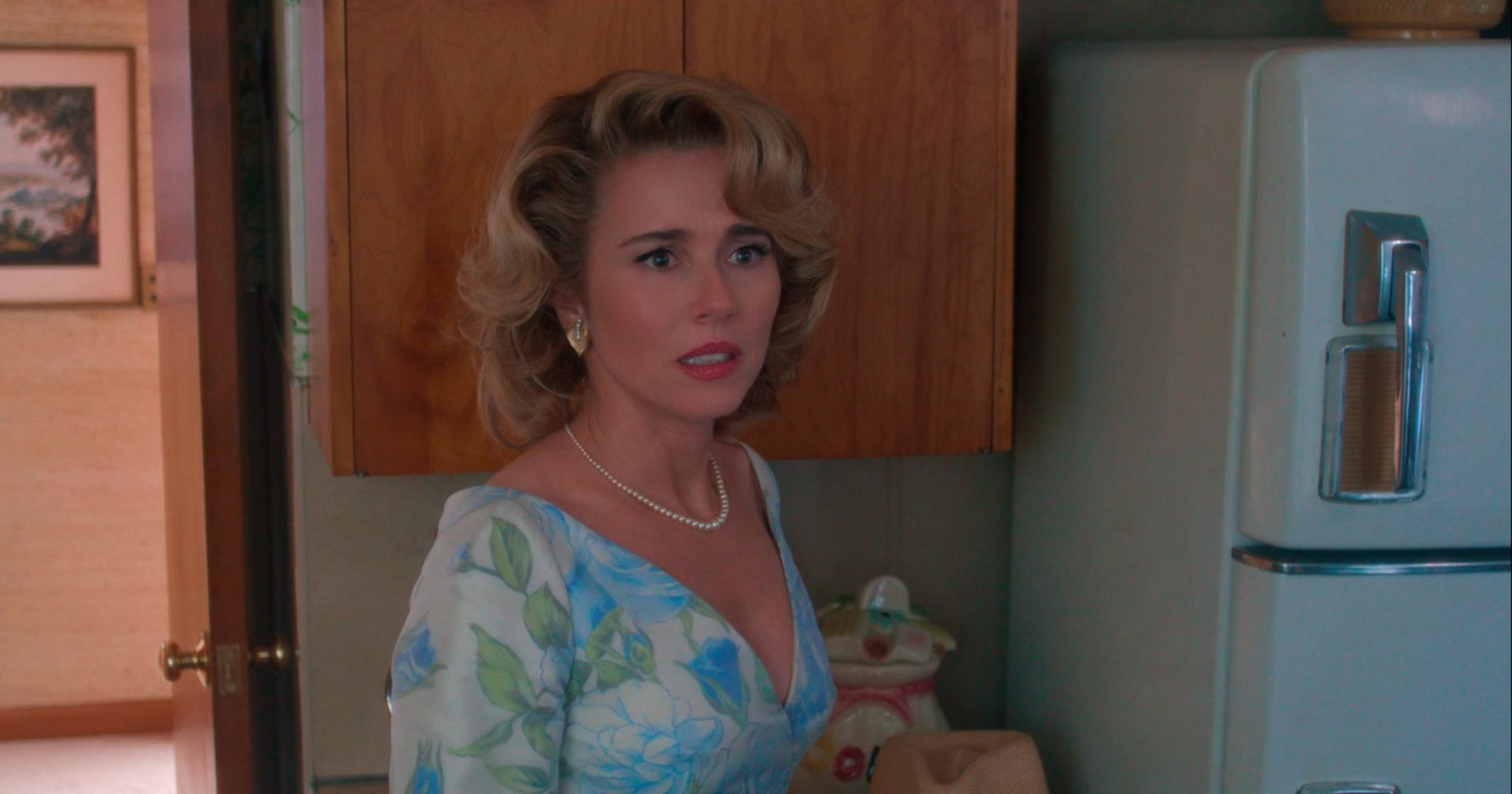 Blonde woman in a blue floral dress and pearl necklace standing in a retro kitchen next to a vintage refrigerator, looking distressed.
