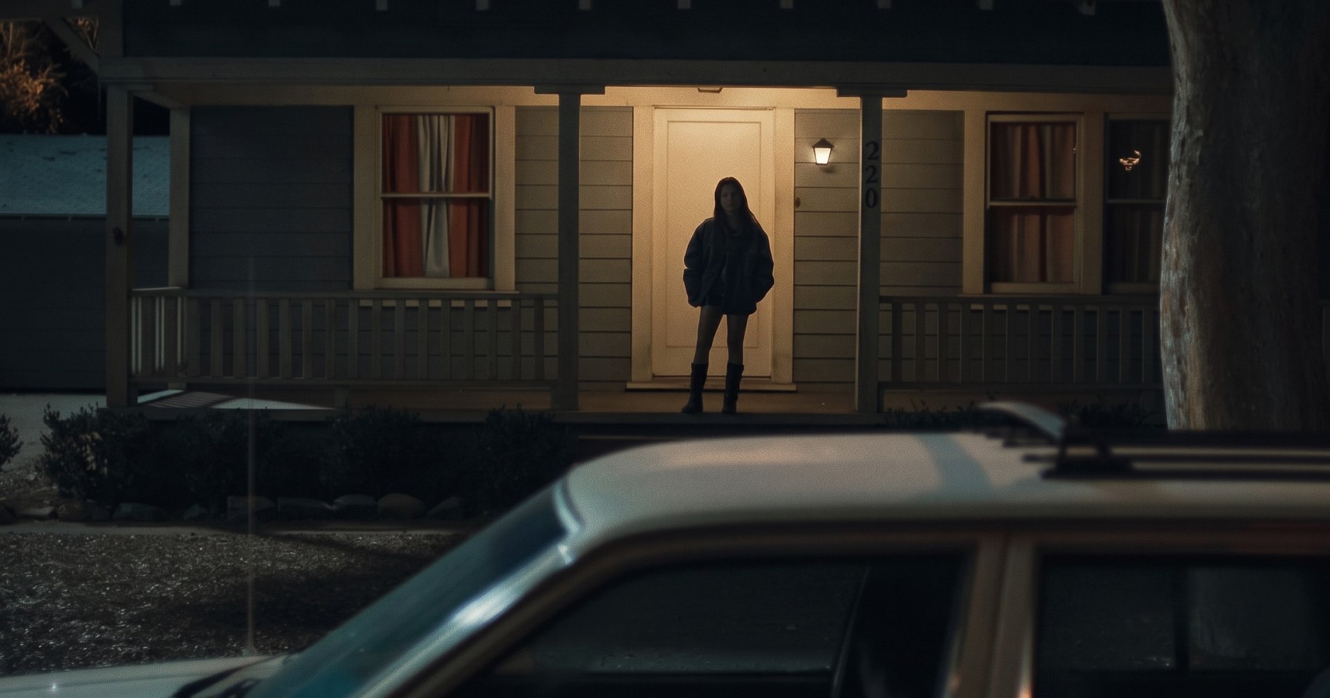 A silhouetted woman stands in front of a lit doorway of a dark suburban house at night, with a car in the foreground.