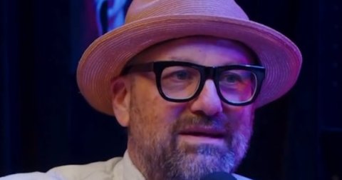 A bearded man wearing a tan fedora and black-rimmed glasses sits in front of a microphone in a dimly lit studio.
