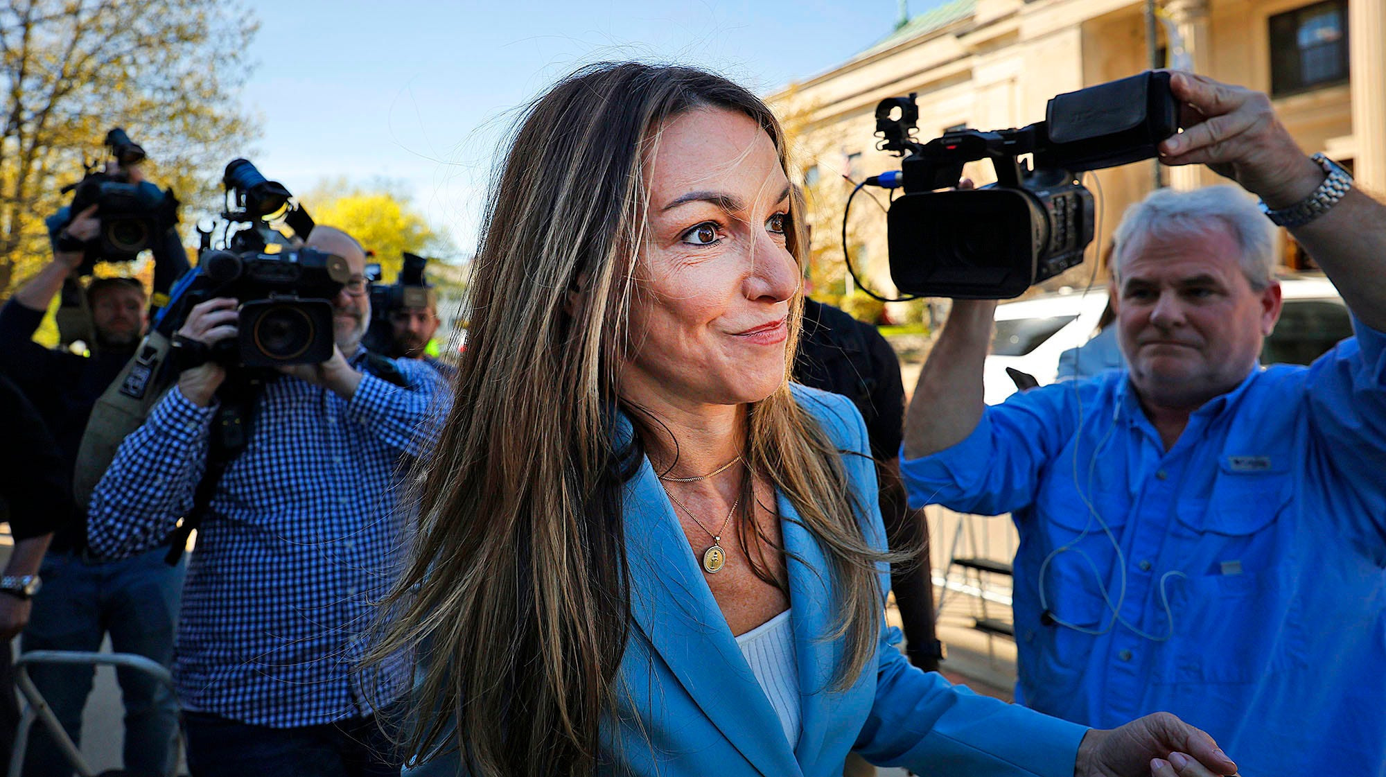 Woman with long brown hair in a light blue blazer walks past TV cameras outside a courthouse.
