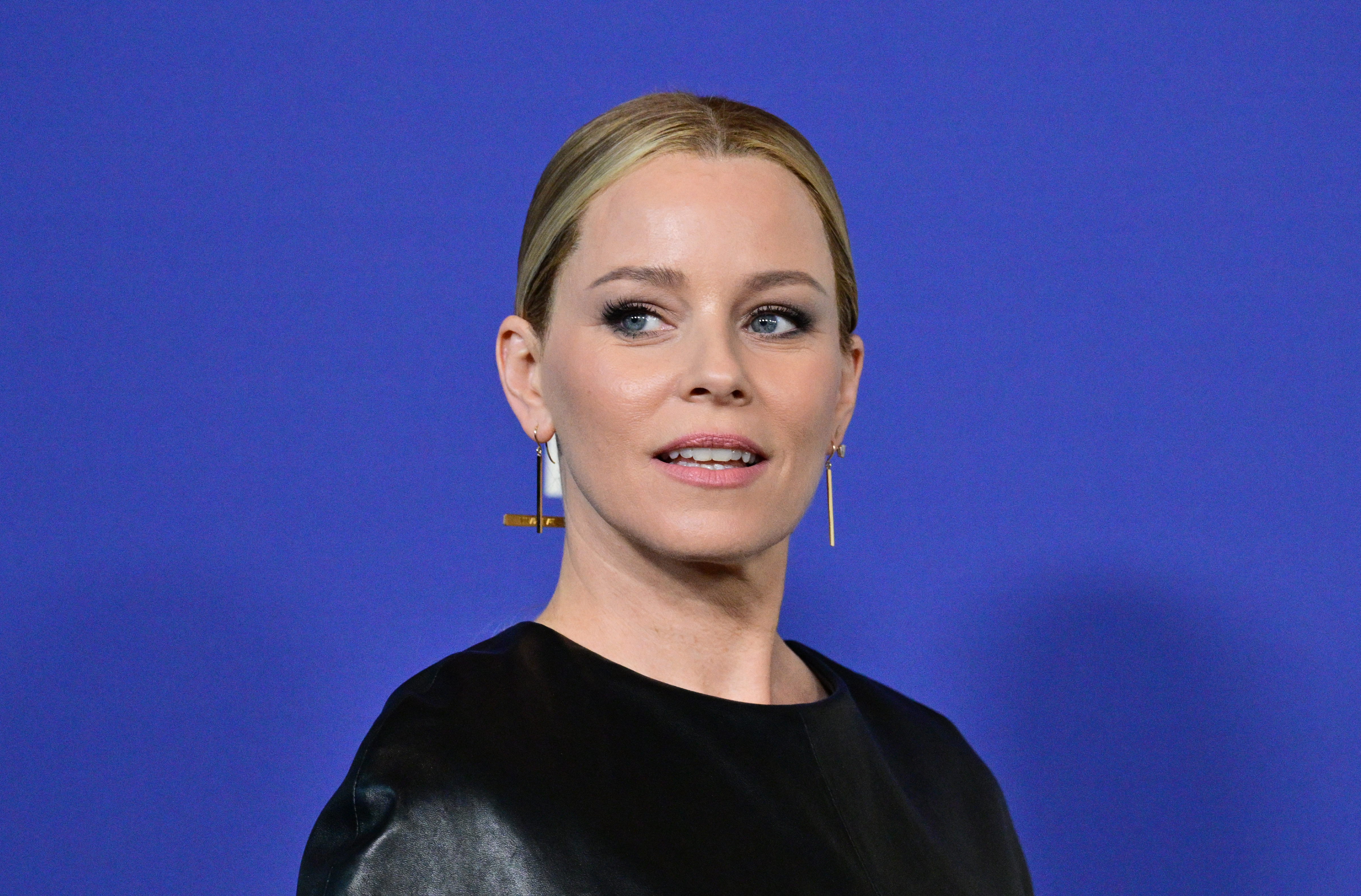 Blonde woman with slicked-back hair wearing gold bar earrings and a black outfit posed against a blue backdrop.
