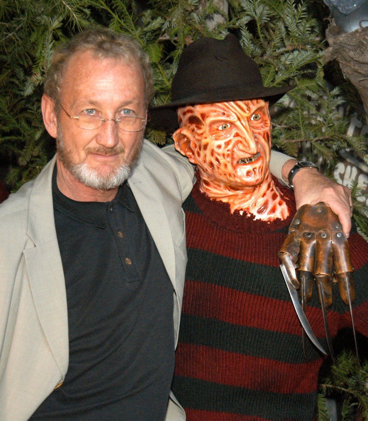 Actor Robert Englund standing next to a Freddy Krueger wax figure wearing a striped sweater, black hat, and bladed glove.