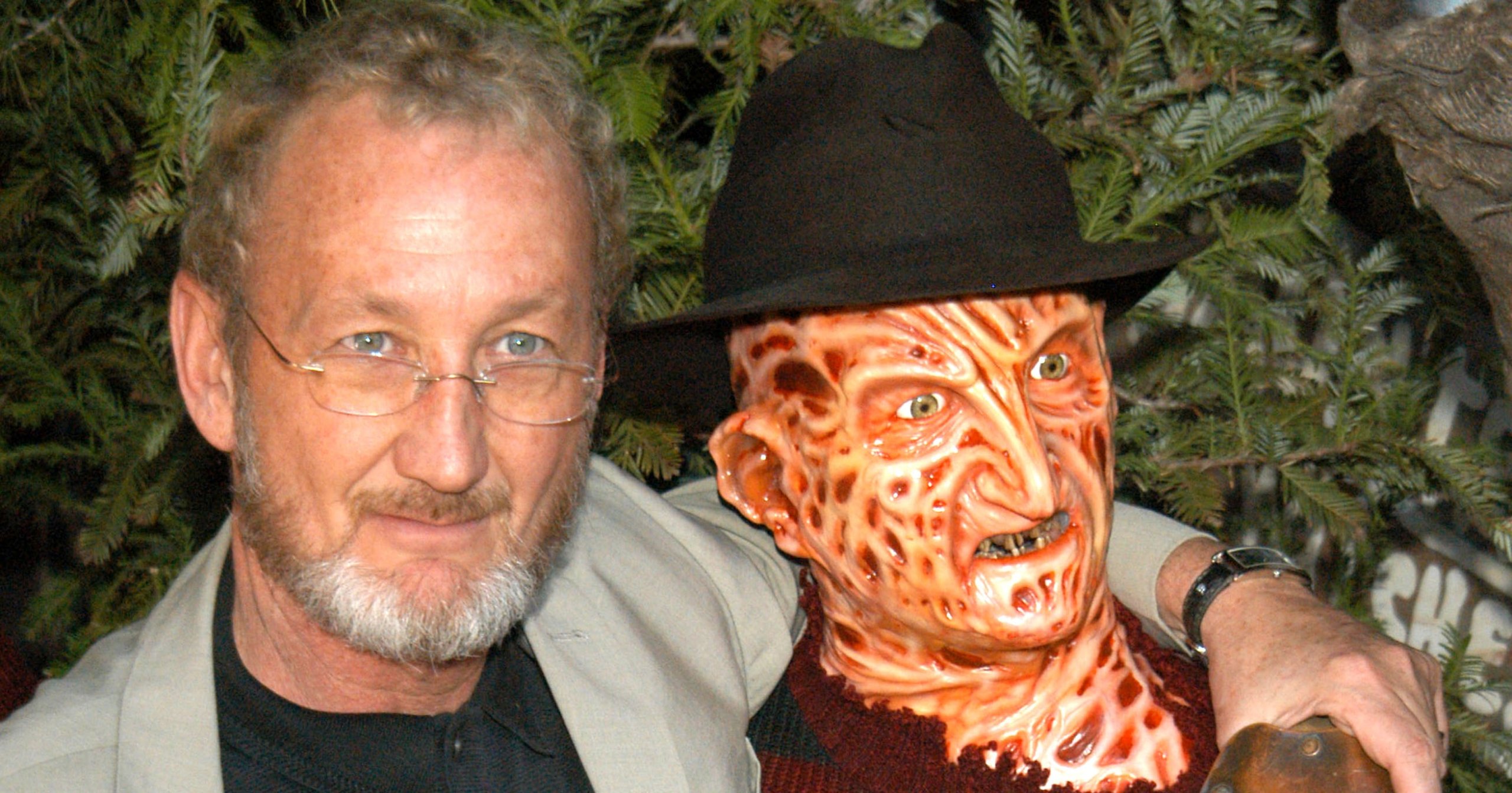 A man with glasses and a goatee stands arm-in-arm with a Freddy Krueger wax figure wearing a striped sweater, fedora, and bladed glove.