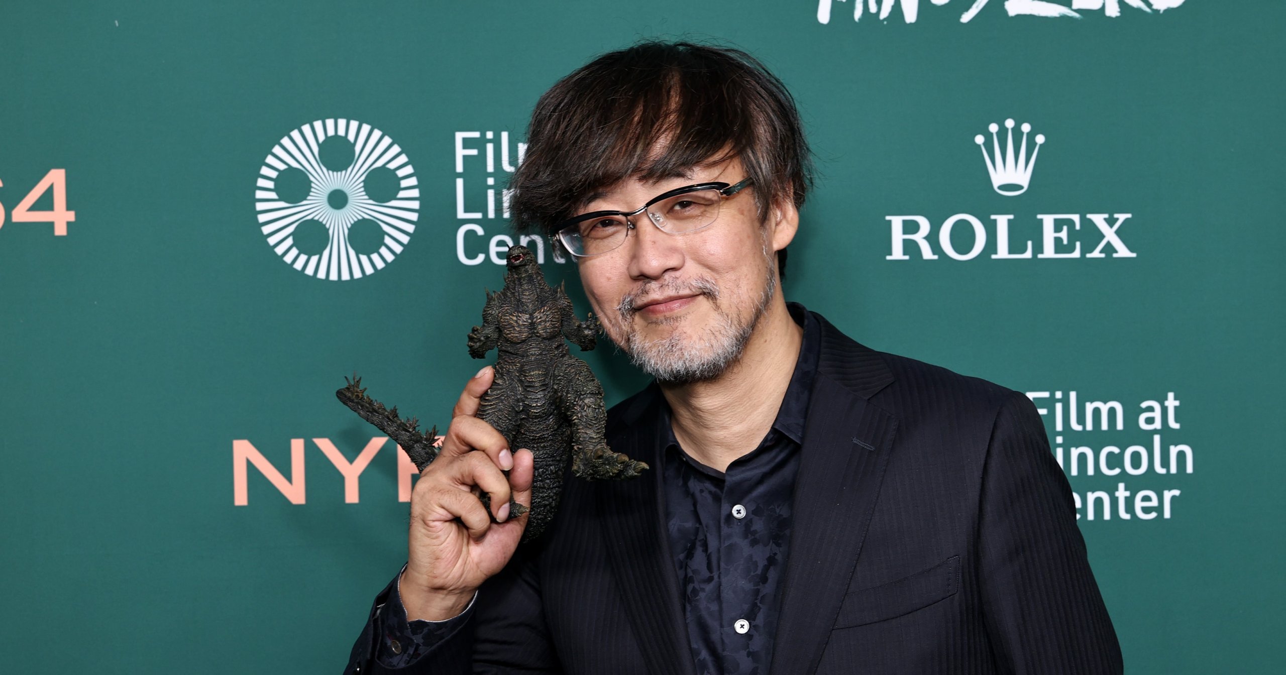 Man in dark suit holding a small Godzilla figurine up near his face, smiling, in front of a green step-and-repeat backdrop for the New York Film Festival.