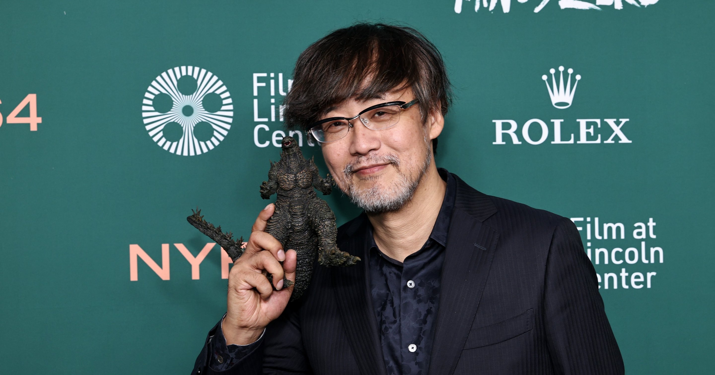 Man in dark suit holding a small Godzilla figurine up near his face, smiling, in front of a green step-and-repeat backdrop for the New York Film Festival.
