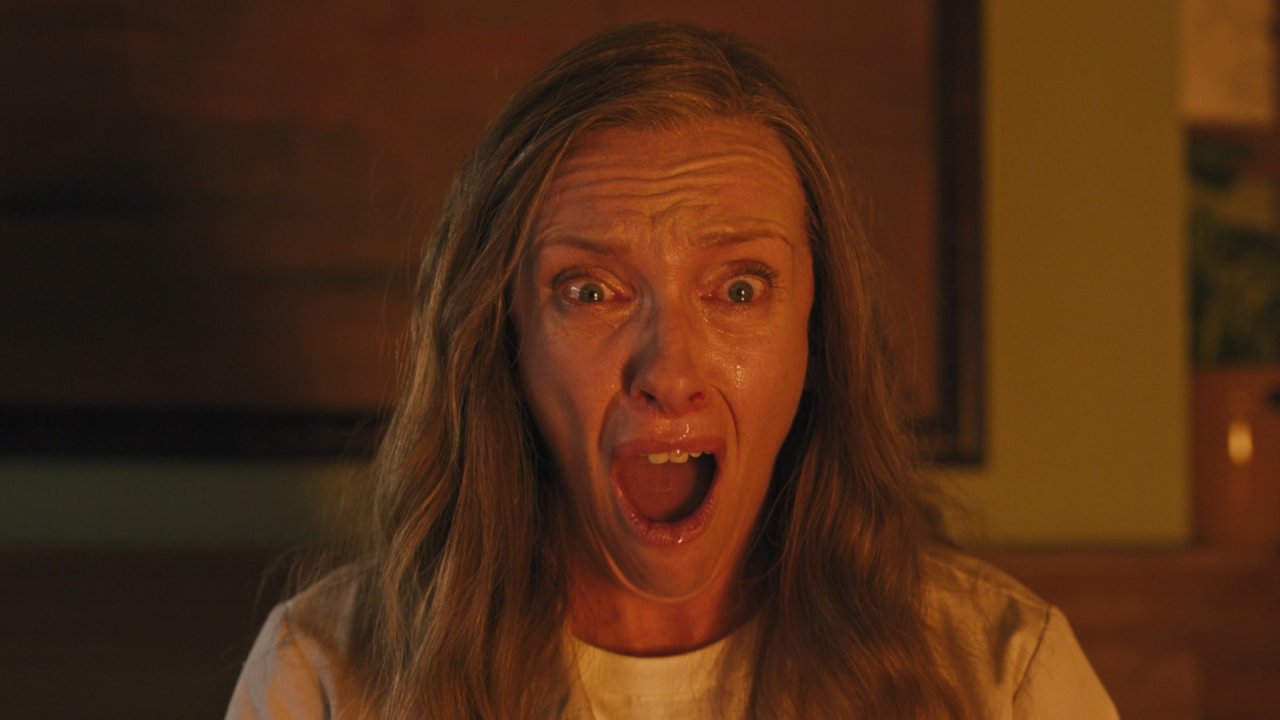 Close-up of a woman with long hair screaming with wide, bloodshot eyes and an open mouth, tears on her face, lit by orange light.