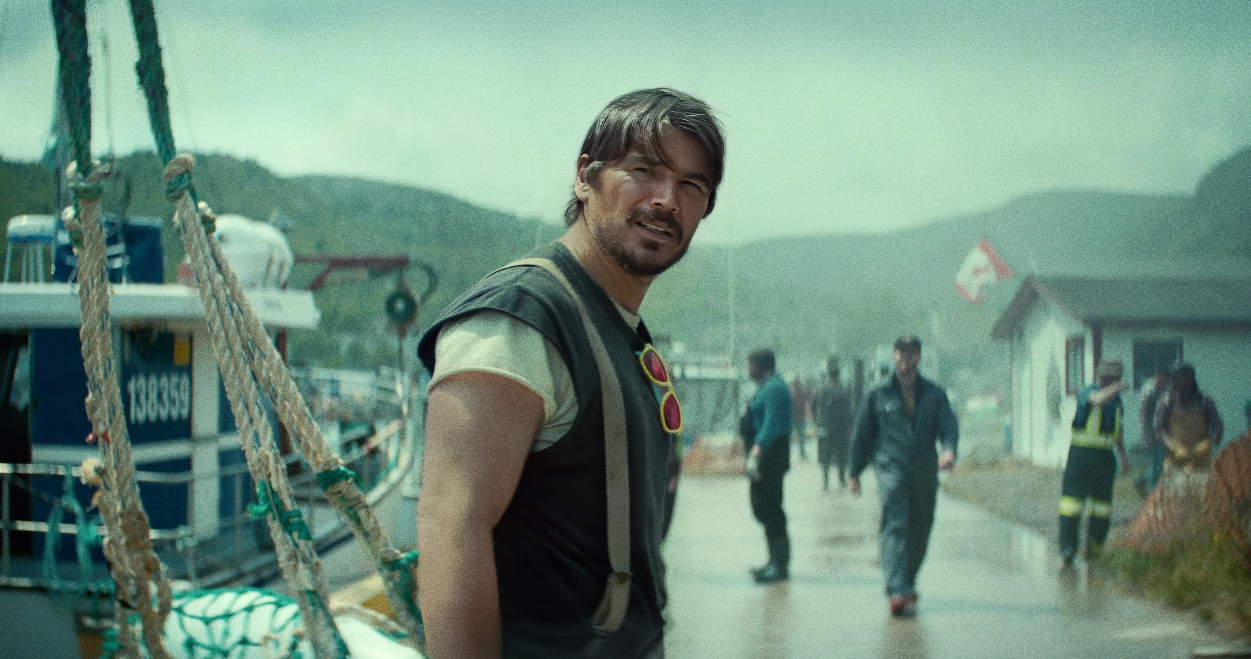 Man in a vest with suspenders and red sunglasses hanging from his collar stands on a rainy fishing dock beside a moored boat.