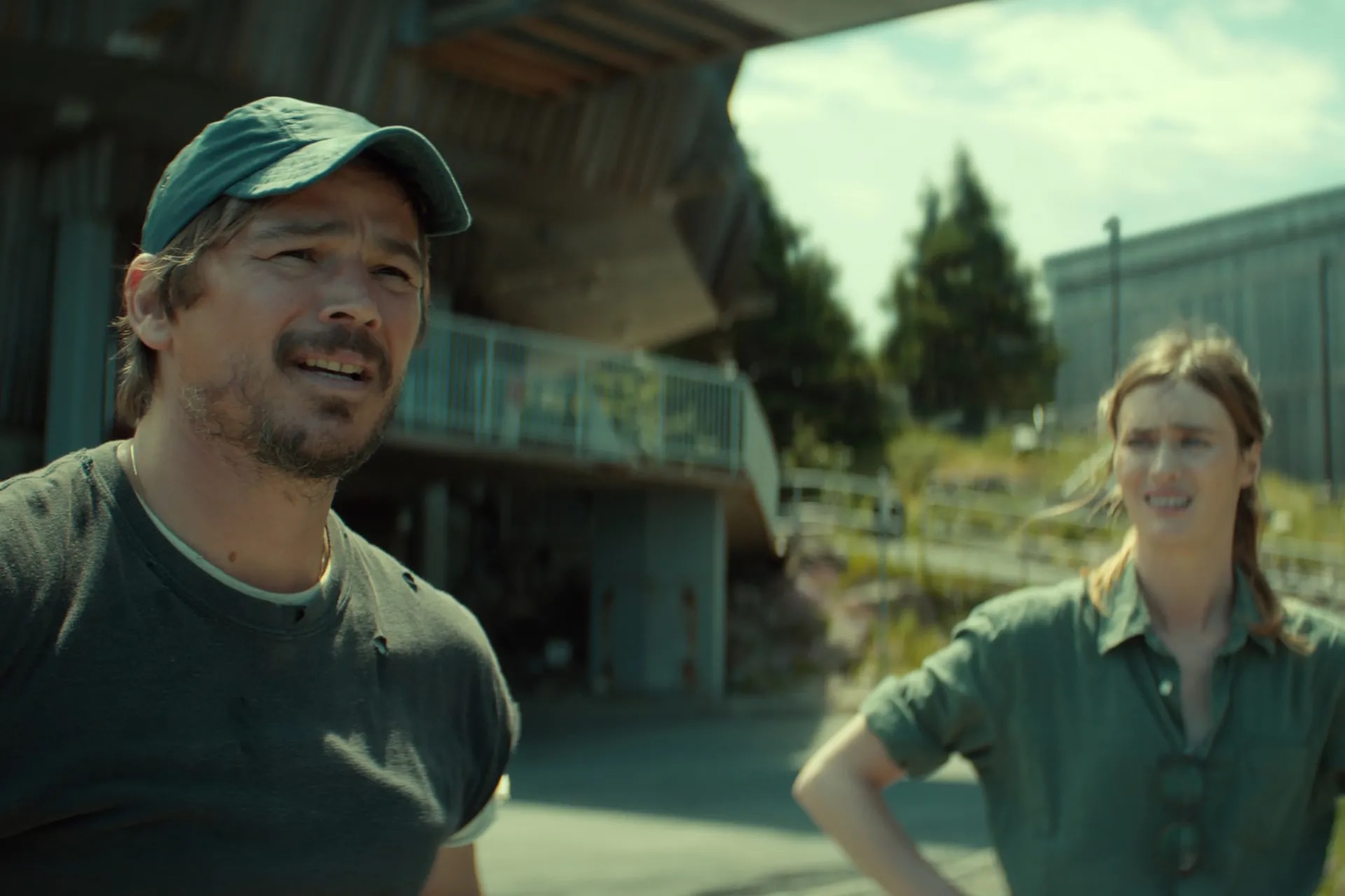 A man in a green cap and worn t-shirt smiles to the side while a woman in a green shirt stands behind him outside a weathered building.