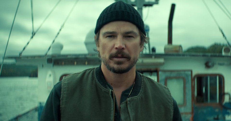 Man in black beanie and olive vest standing on a fishing boat deck with overcast skies, from Netflix's Below.