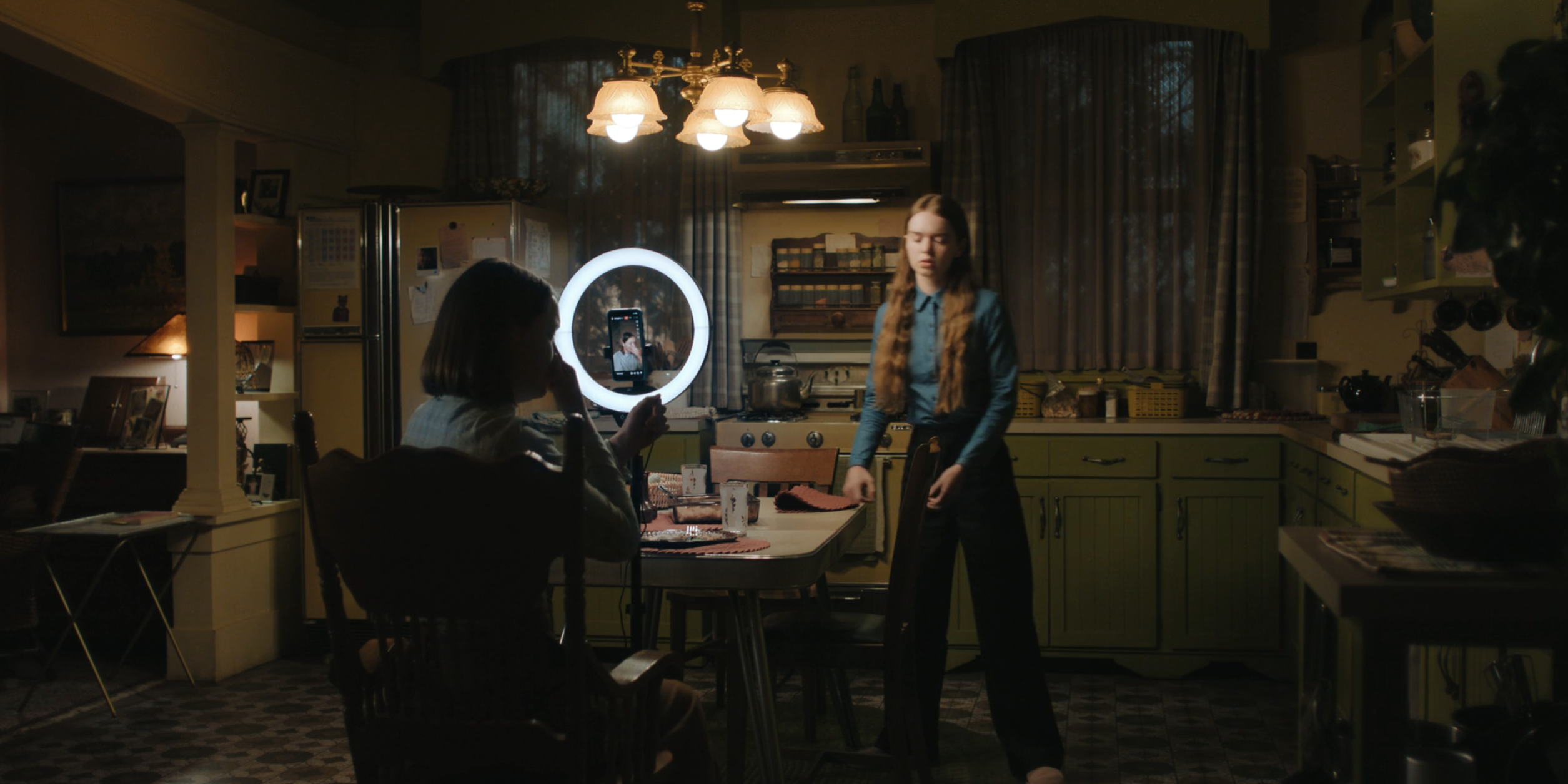 Wide shot of a vintage-style kitchen: a woman sits at a table facing a ring light and phone, while a teenage girl with long red hair stands near the stove.