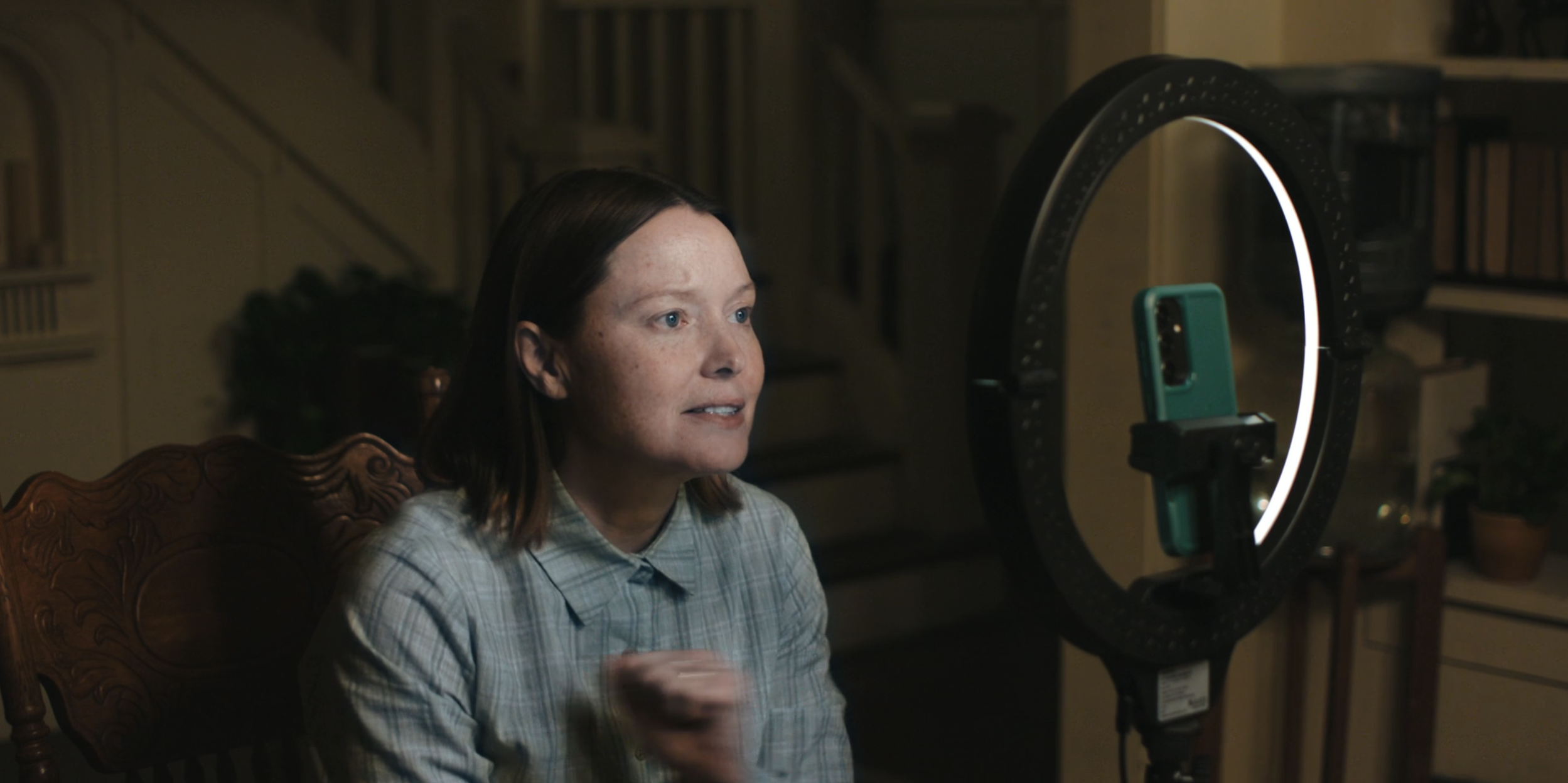 A woman with short brown hair speaks toward a smartphone mounted in a ring light on a tripod, seated in a wooden chair in a dimly lit home.