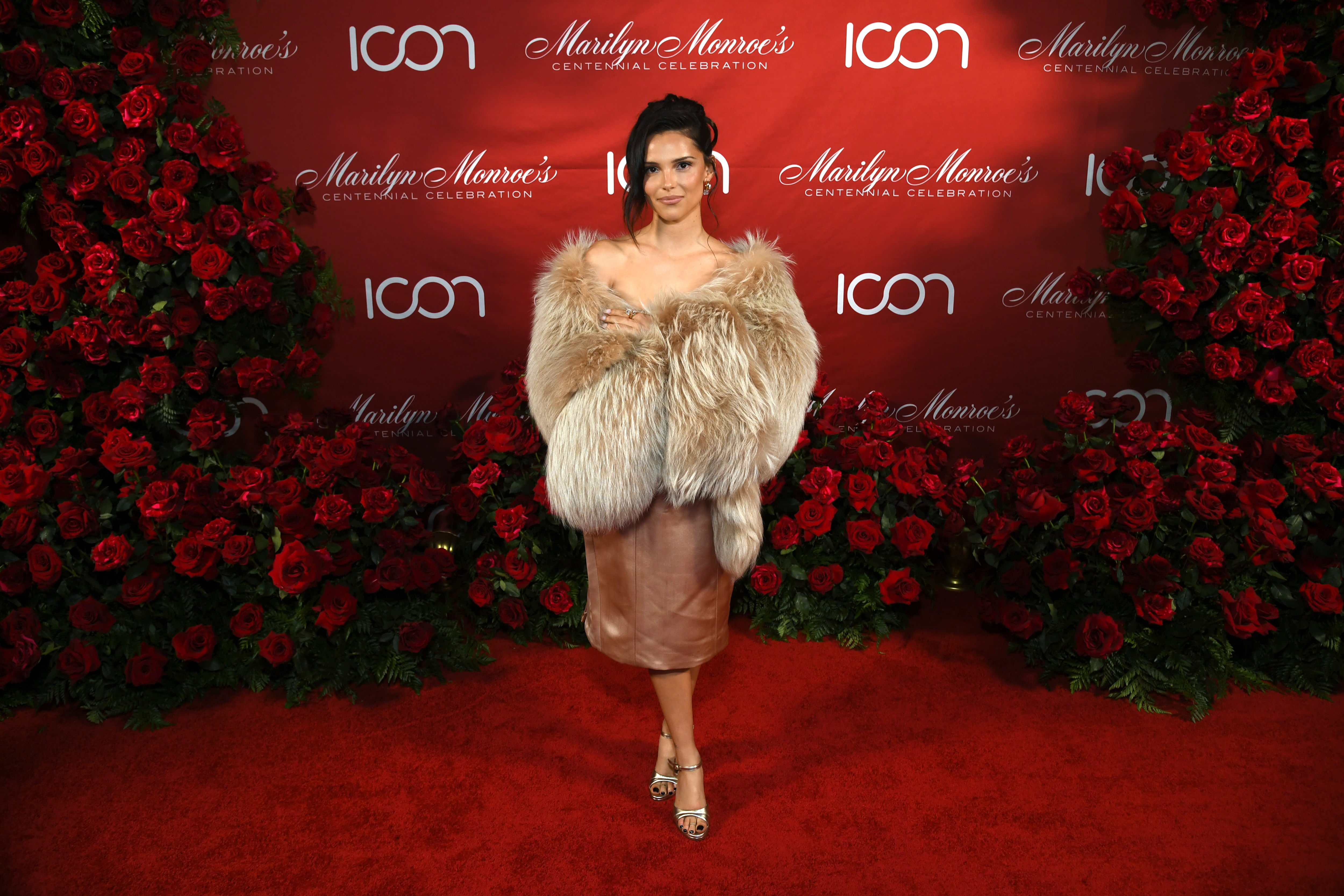 Inde Navarrette in a fur stole and satin skirt poses on the red carpet at Marilyn Monroe's Centennial Celebration, surrounded by red roses.