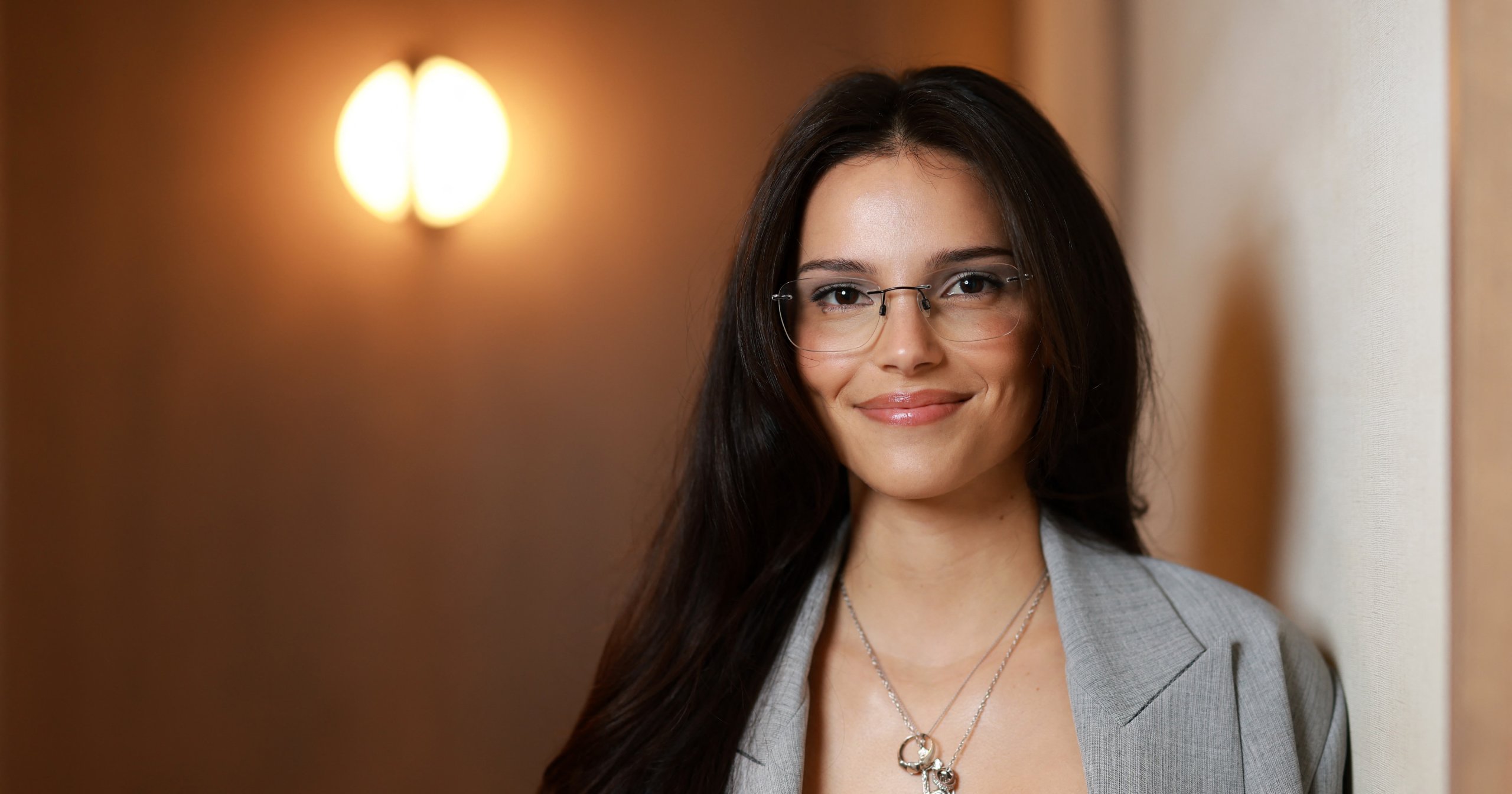 Actress Inde Navarrette smiling in a gray blazer and glasses, posing indoors at TIFF 2026.
