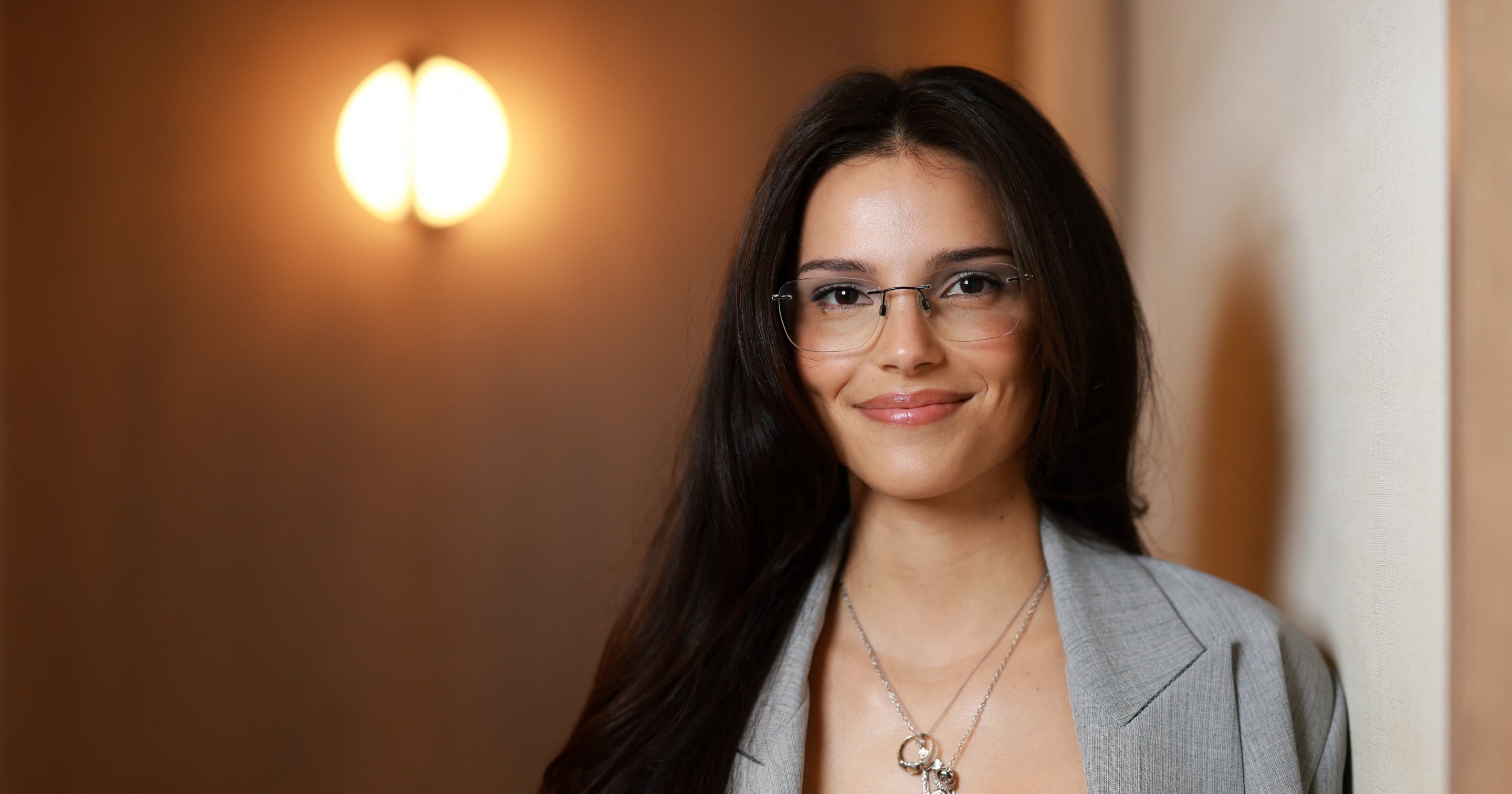 Actress Inde Navarrette smiling in a gray blazer and glasses, posing indoors at TIFF 2026.