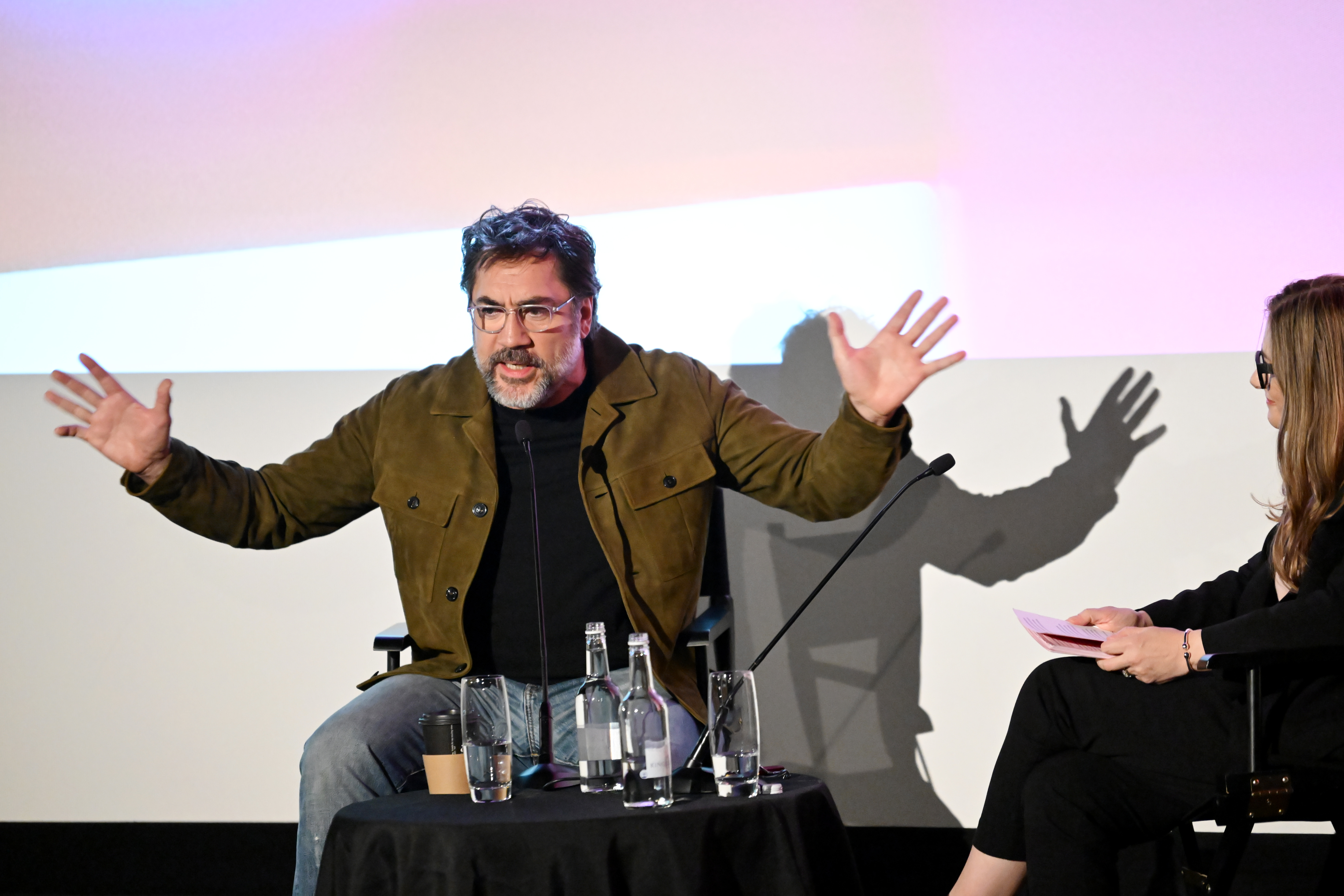 A bearded man in glasses and an olive jacket sits on stage with both hands raised, speaking into a microphone, beside a woman holding papers.