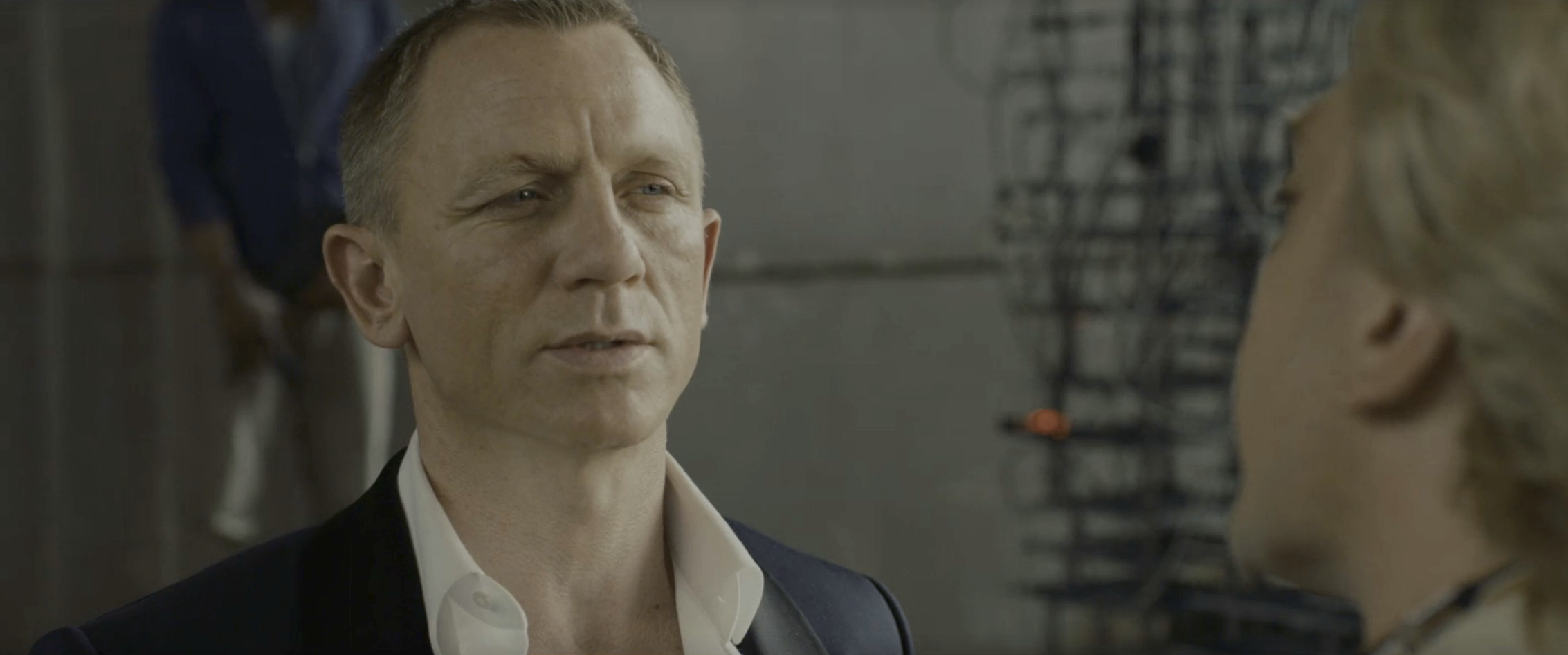A short-haired man in a navy blazer with an open white shirt looks intently to the side, with a blurred figure in the foreground.