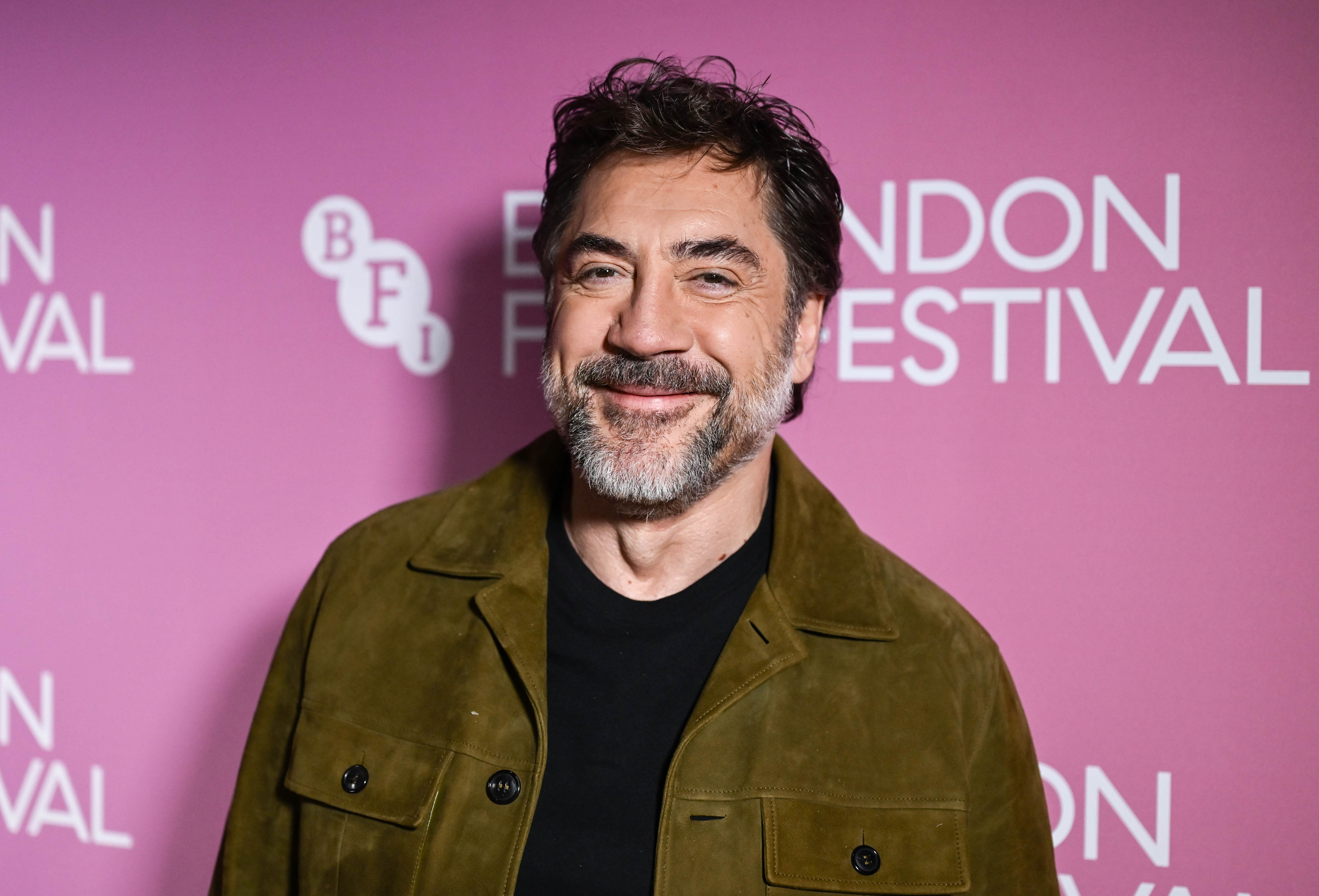 A smiling bearded man in an olive suede jacket stands in front of a pink BFI London Film Festival step-and-repeat backdrop.
