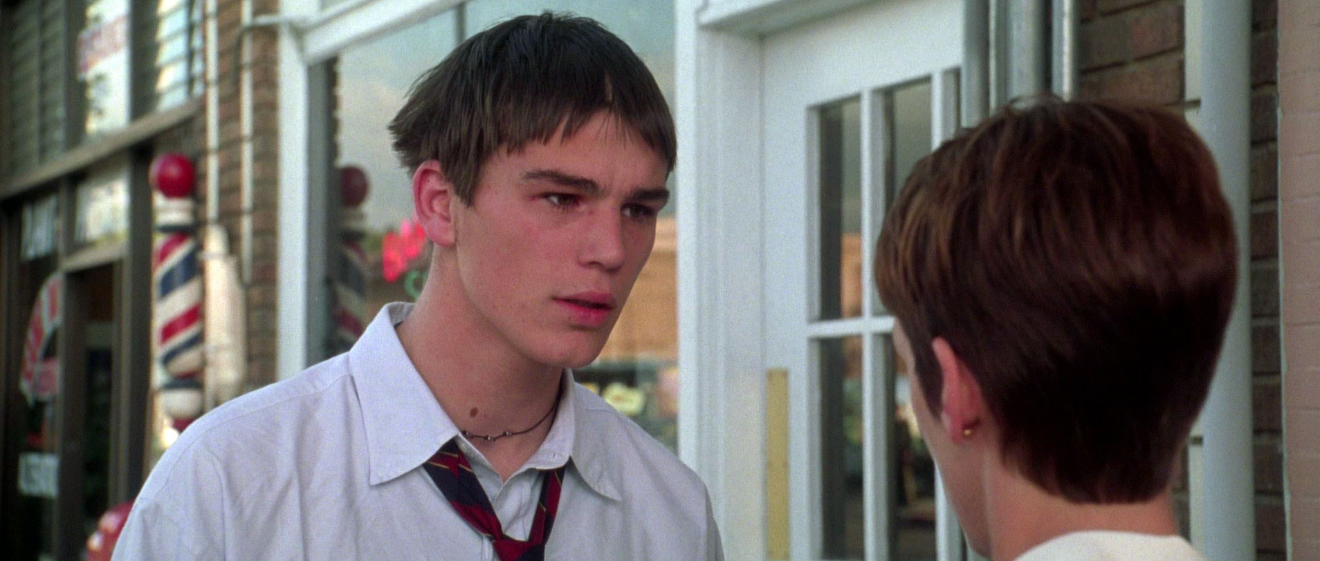 Young man with choppy dark bangs in a white shirt and loosened striped tie facing another person, a barber pole visible behind them.