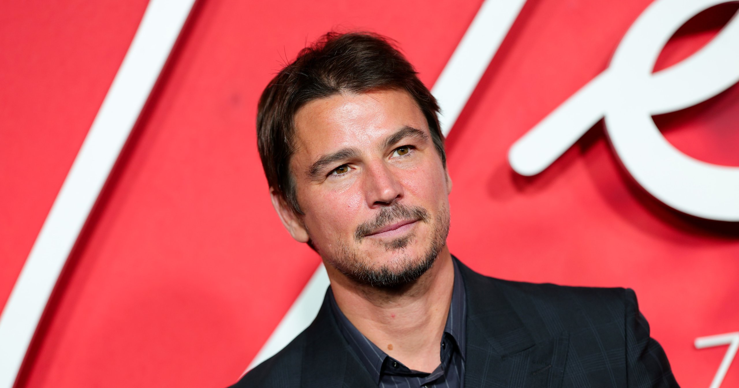 Josh Hartnett in a dark suit and striped shirt posing in front of a red step-and-repeat backdrop.
