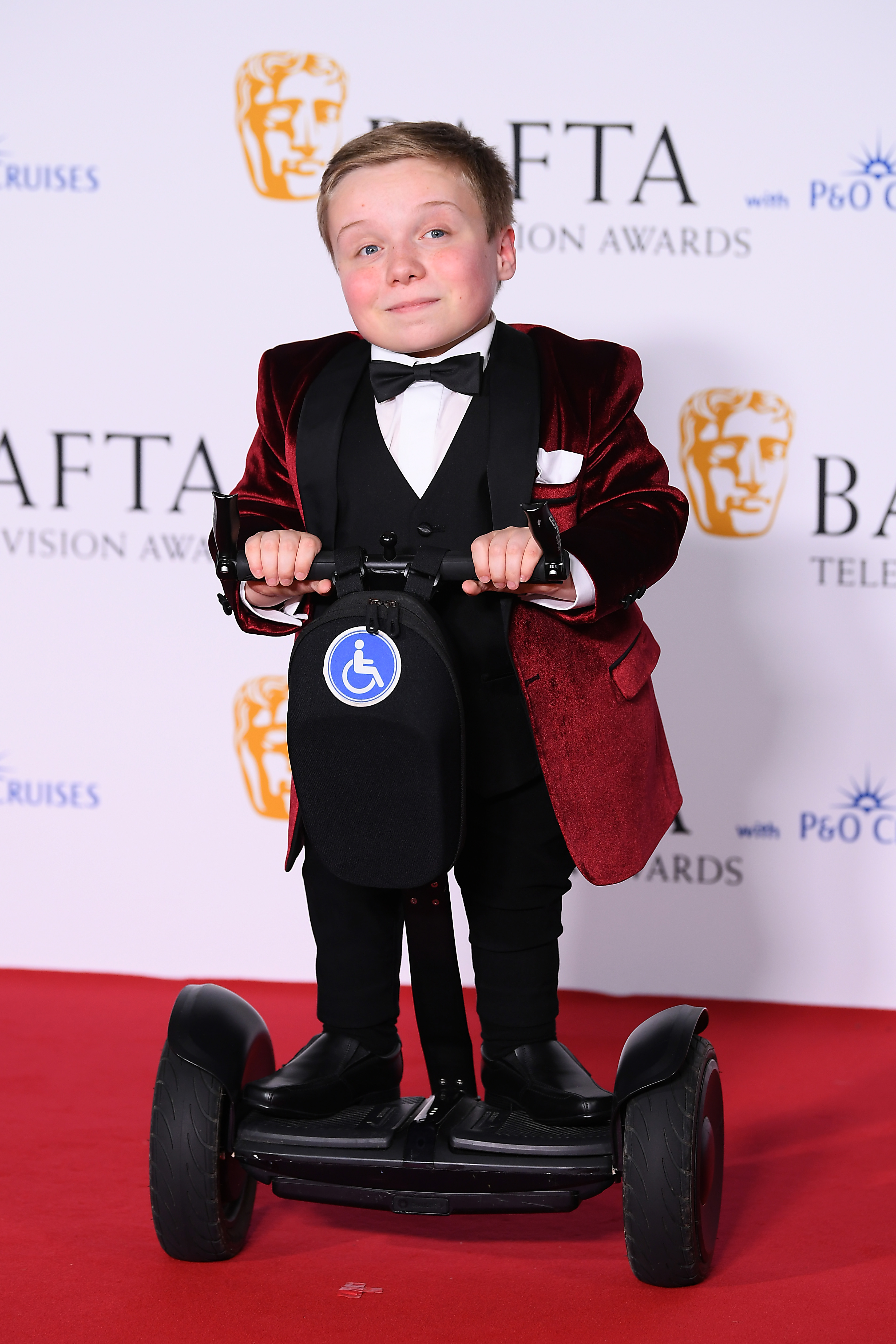 Young actor Lenny Rush in a red velvet tuxedo on a mobility scooter at the BAFTA Television Awards red carpet, London.