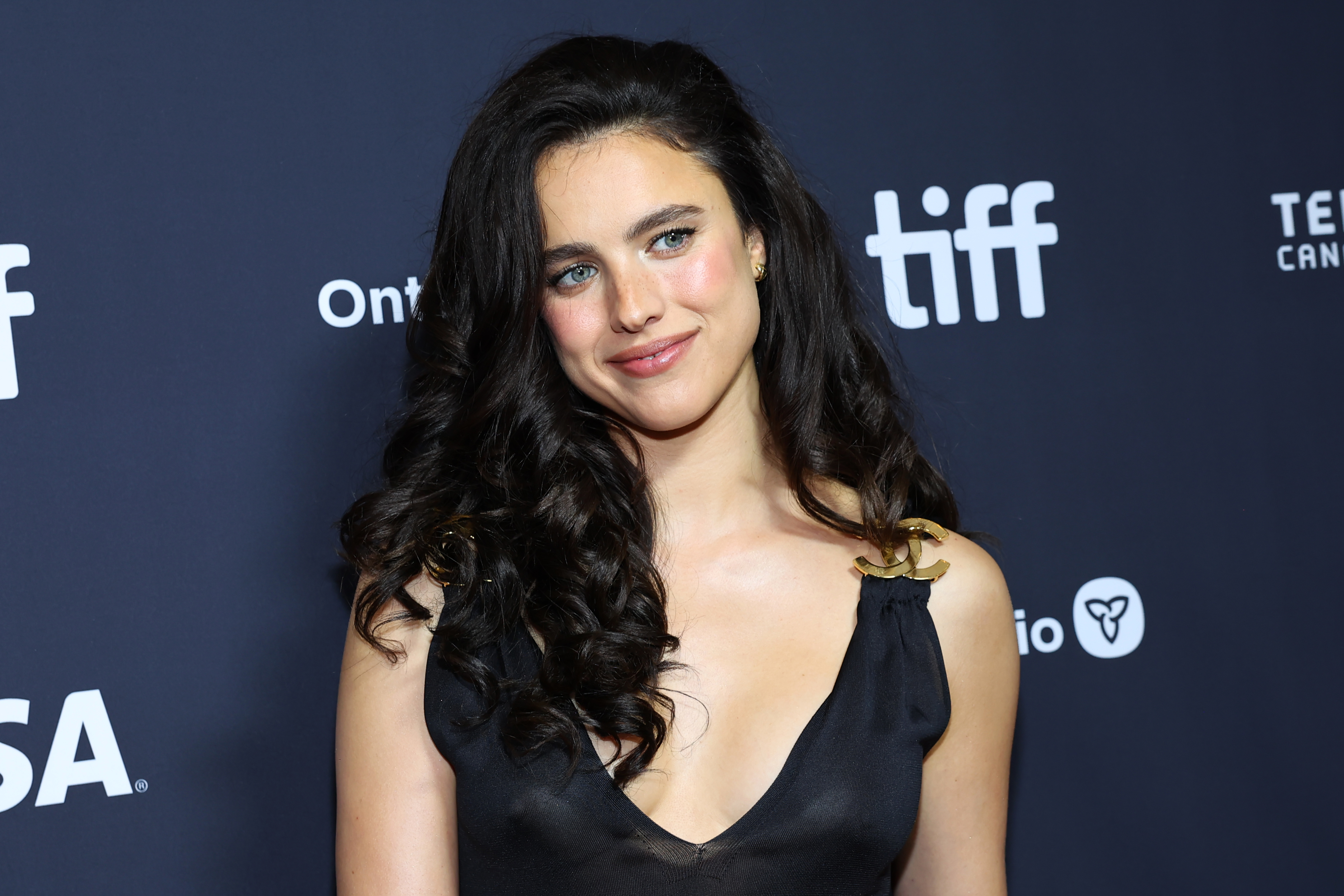 Margaret Qualley in a black dress with gold Chanel hardware, smiling at the TIFF photo call backdrop.