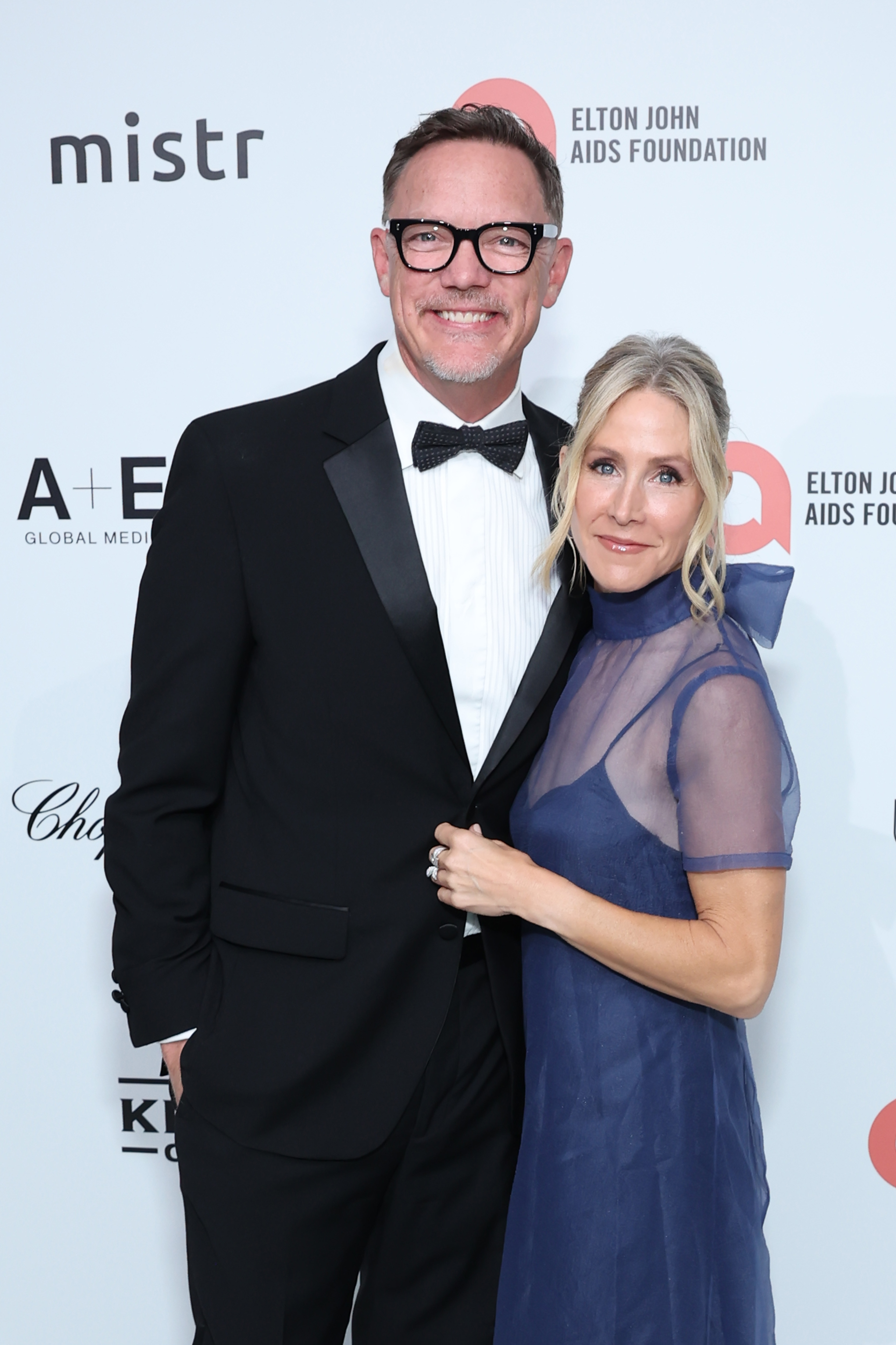 Matthew Lillard in a black tuxedo and bow tie stands with Heather Lillard in a blue sheer dress at the Elton John AIDS Foundation event.