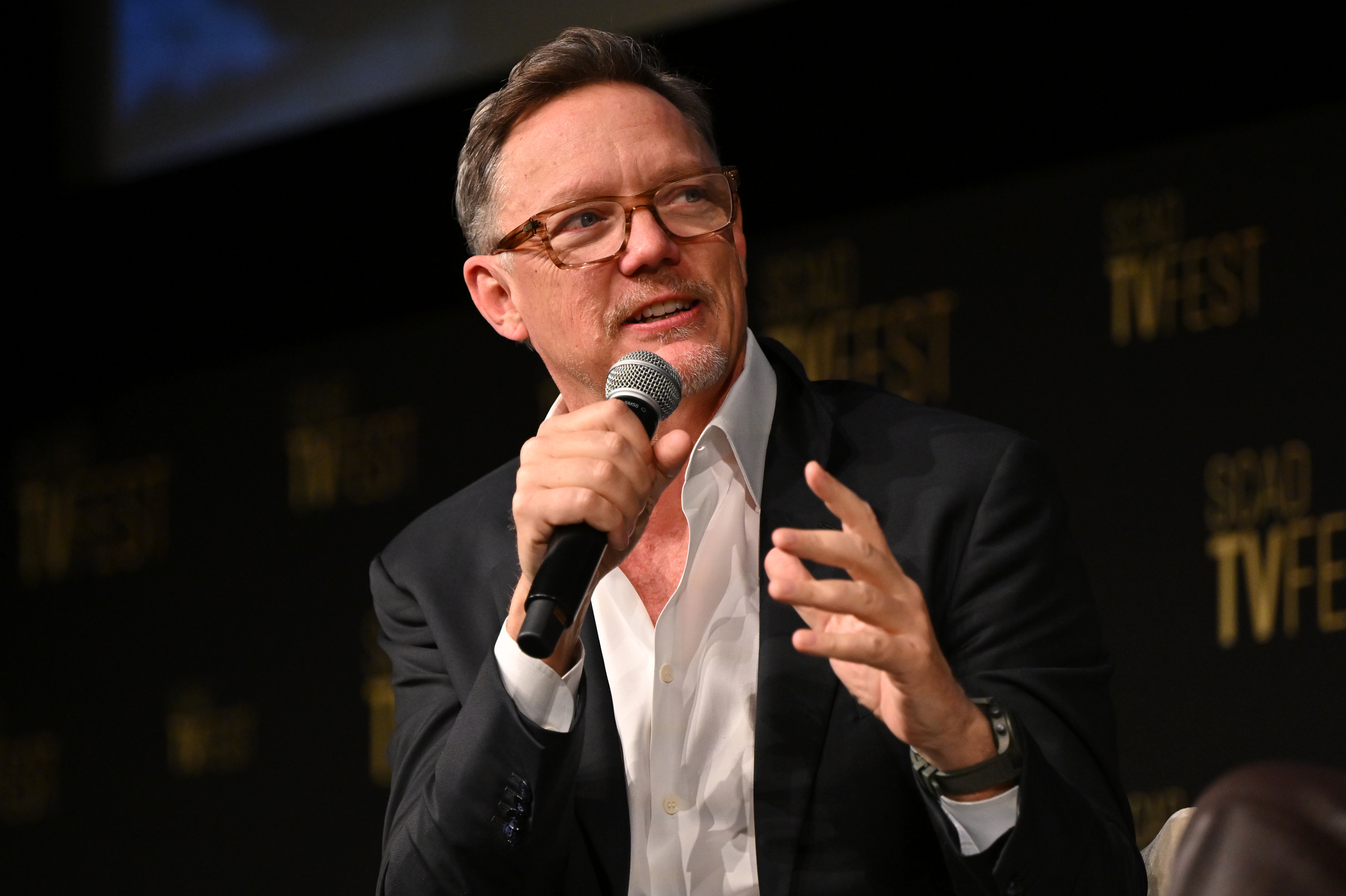 Man with glasses in a dark blazer and white shirt speaking into a handheld microphone at a panel event with a TVfest backdrop.