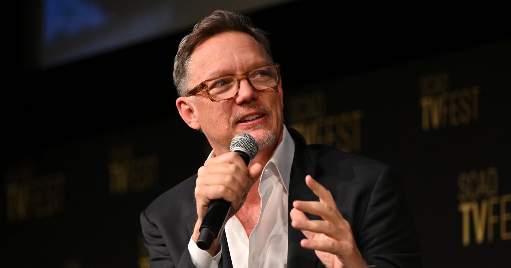 Man with glasses in a dark blazer and white shirt speaks into a handheld microphone at a panel event, gesturing with his other hand, against a black SCAD TVfest backdrop.