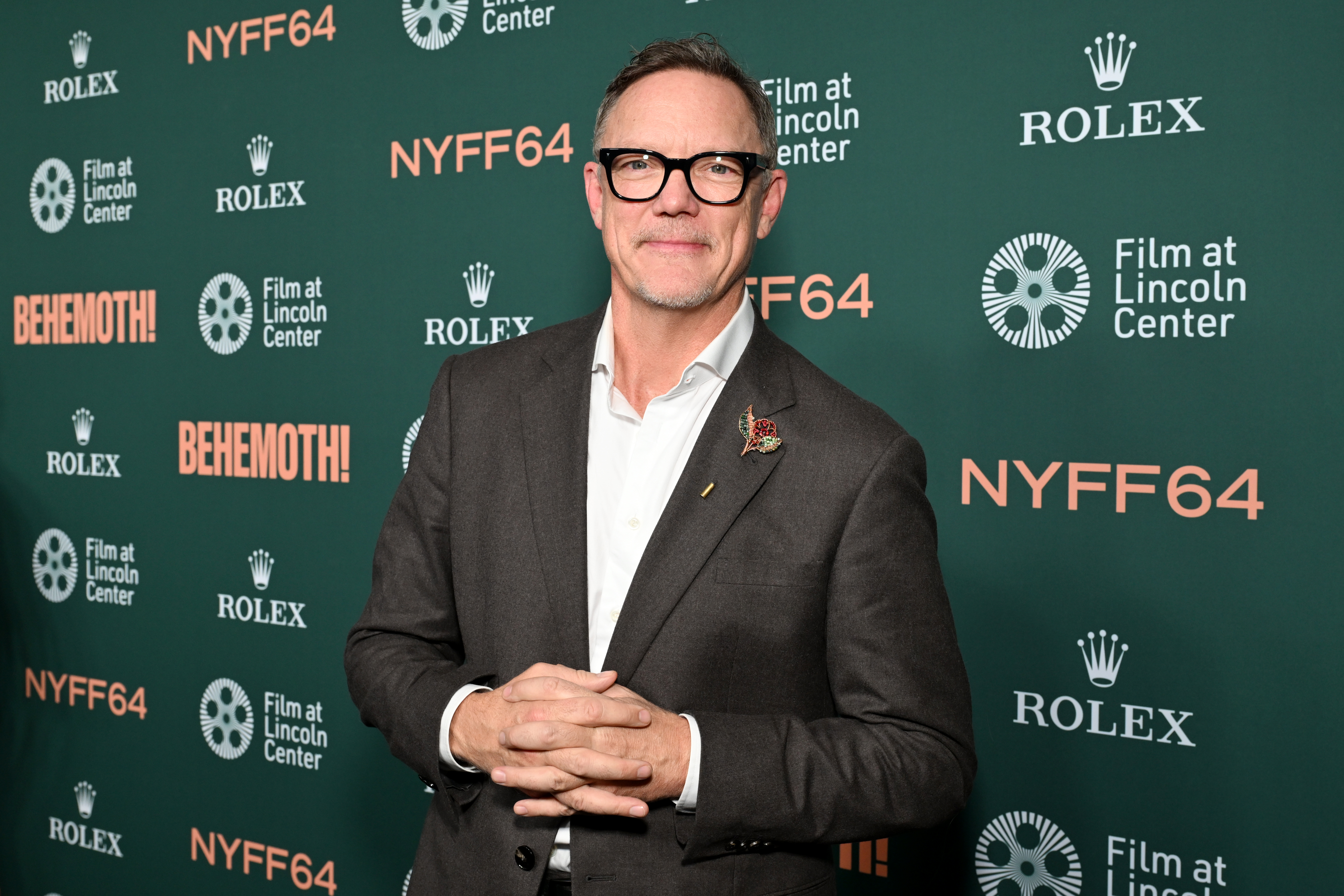 Matthew Lillard in a dark blazer and black-rimmed glasses, hands clasped, standing before a green NYFF64 step-and-repeat backdrop.