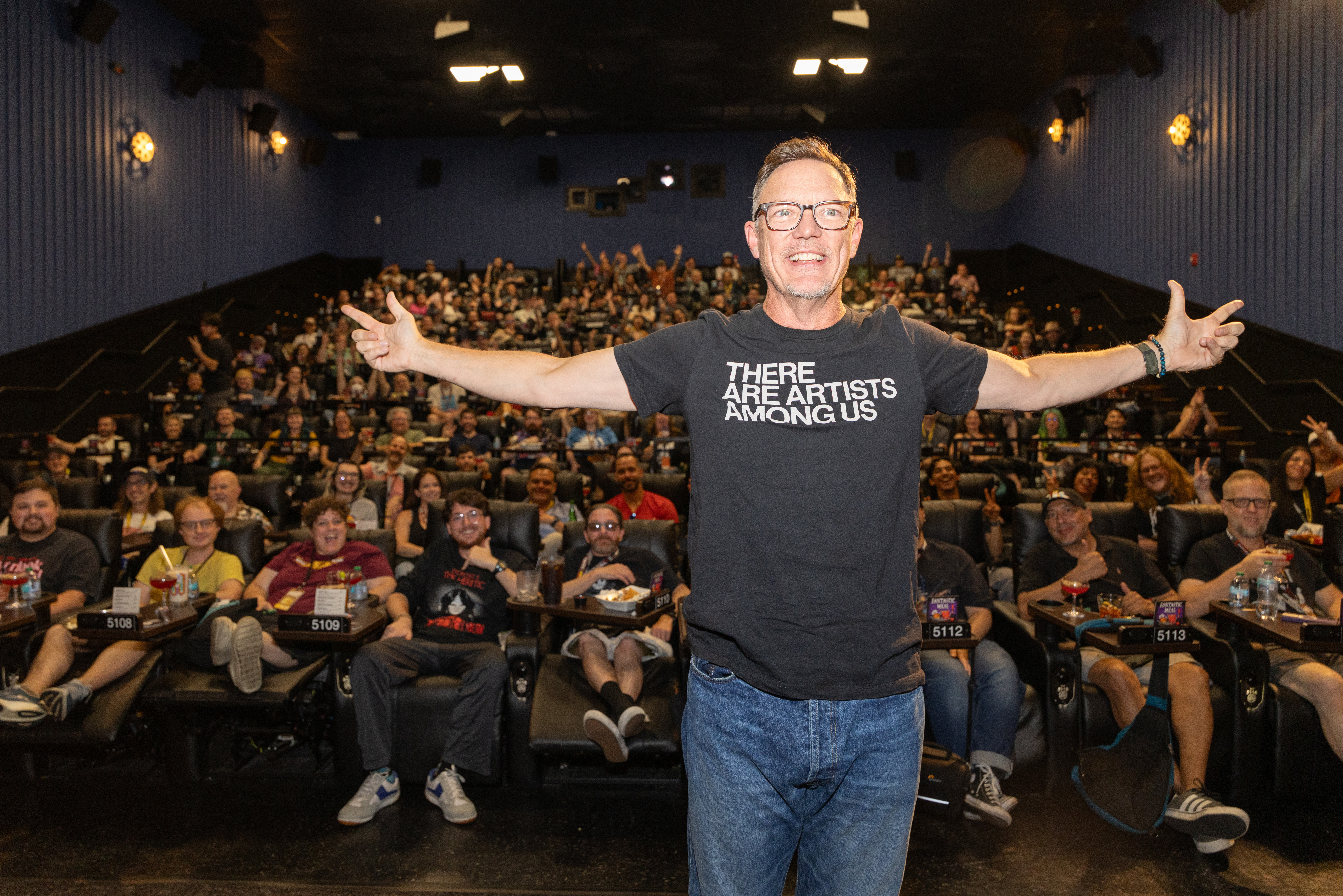 Matthew Lillard in a black 'There Are Artists Among Us' t-shirt stands arms wide in front of a full movie theater audience.