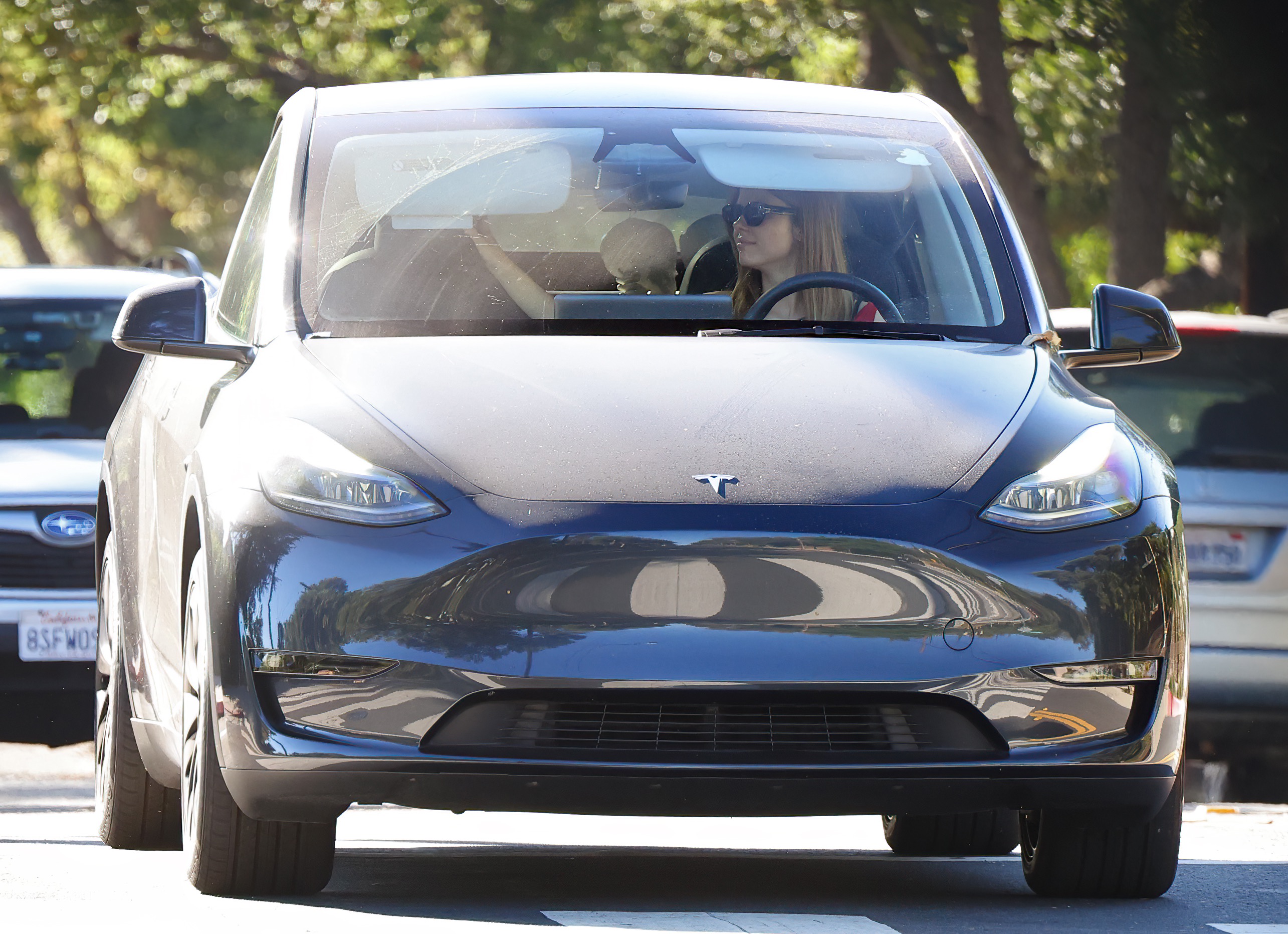 Dark gray Tesla Model Y seen from the front with a woman wearing sunglasses in the driver's seat on a tree-lined street.