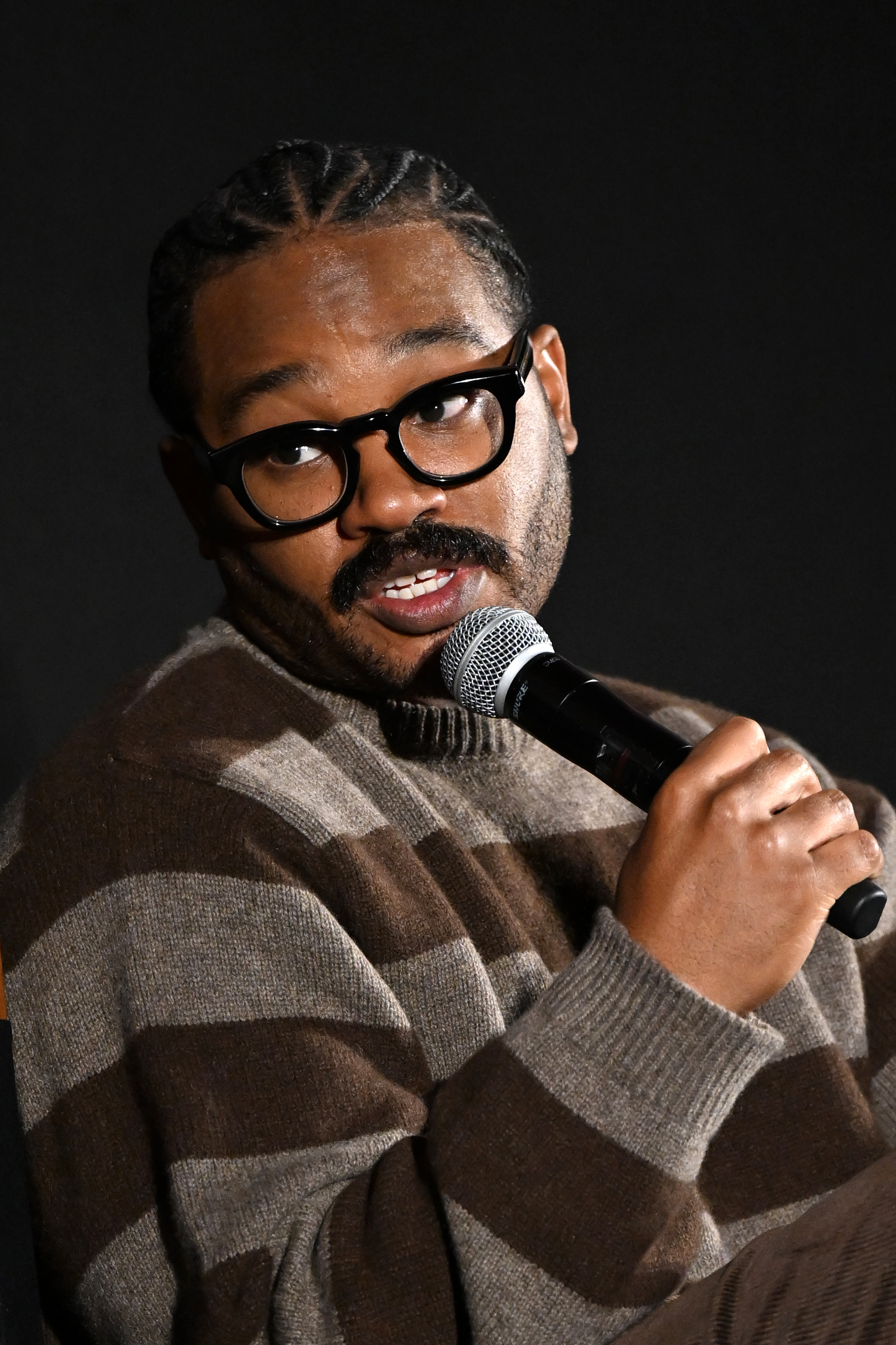 Ryan Coogler holding a microphone and speaking, wearing round black-framed glasses and a brown striped turtleneck sweater, against a dark background.