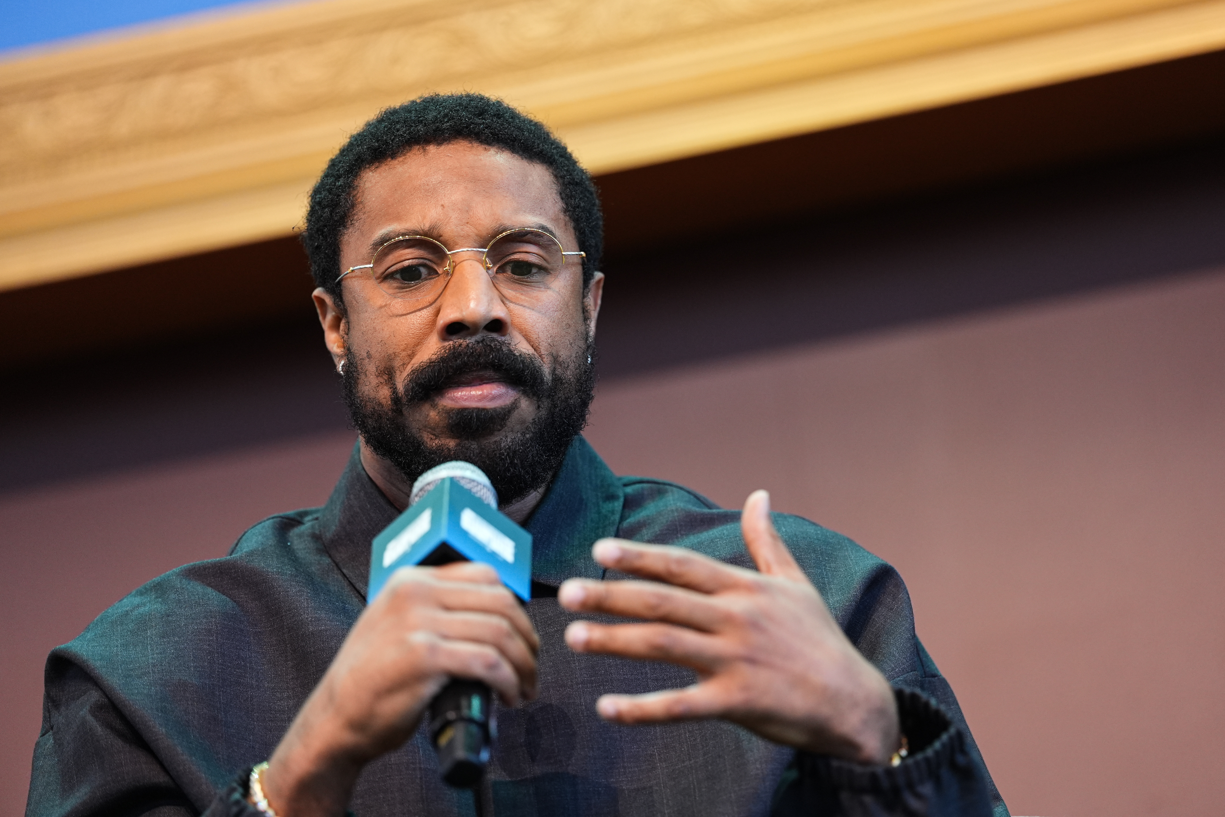 Man with short natural hair, round gold-framed glasses, and a dark jacket speaks into a handheld microphone while gesturing with his other hand.