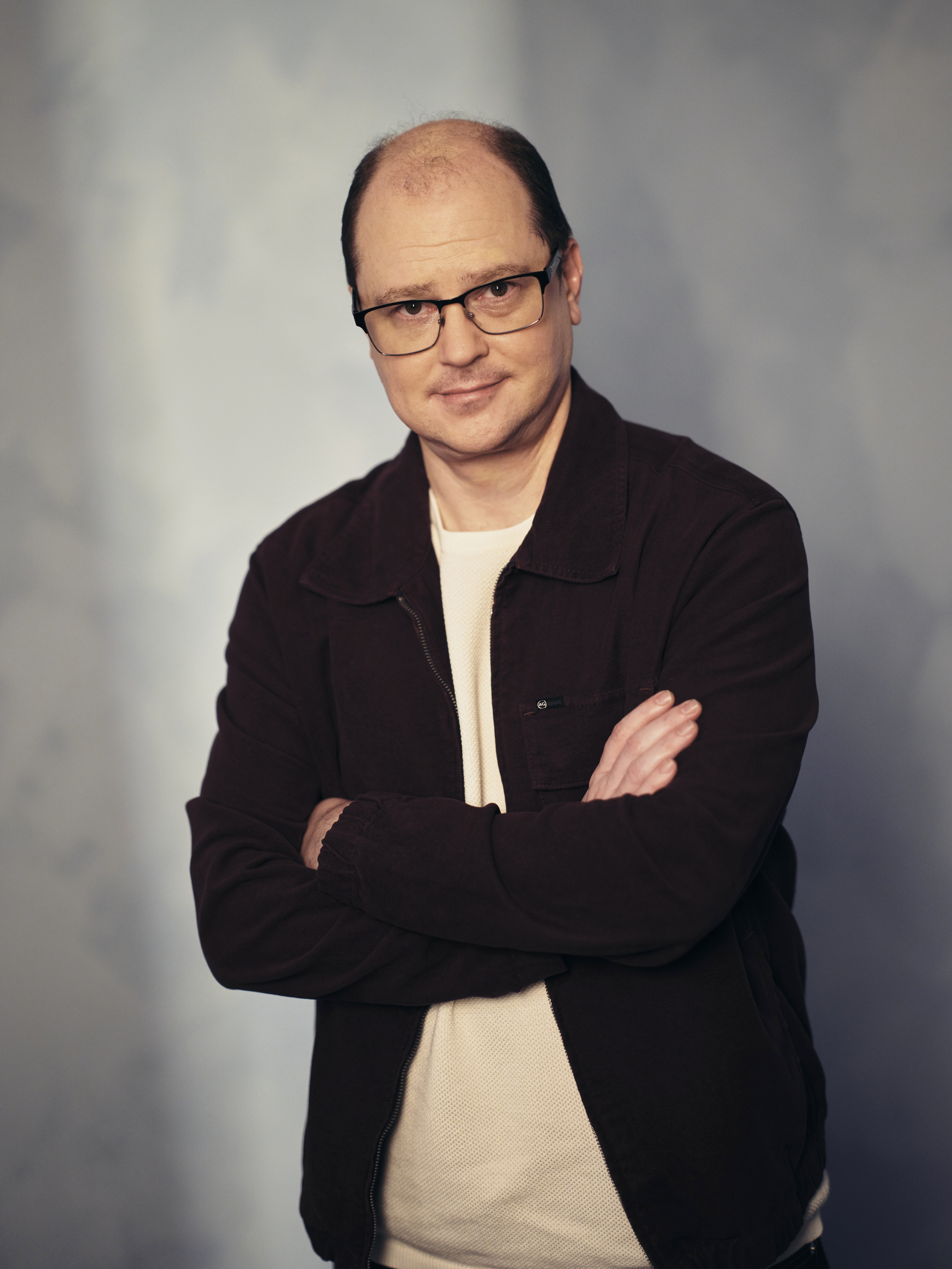 Bald man with glasses in a dark zip-up jacket and white shirt, arms crossed, posing against a grey backdrop.