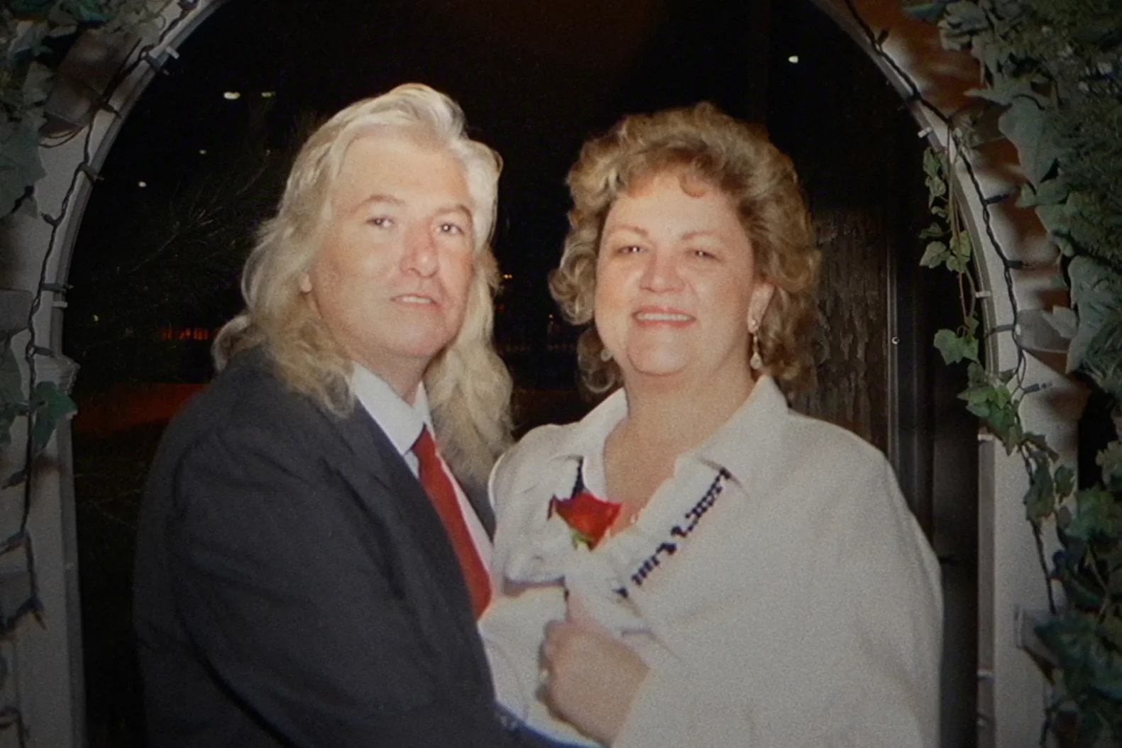 A man with long blond hair in a dark suit and red tie stands with a smiling curly-haired woman in a white top beneath a vine-covered arch.