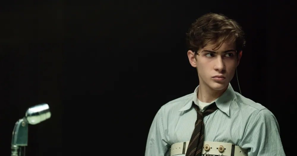 Young teenage boy in a button-up shirt and tie, wearing a white electrode harness with red wires, seated at a table with a vintage microphone, looking tense.