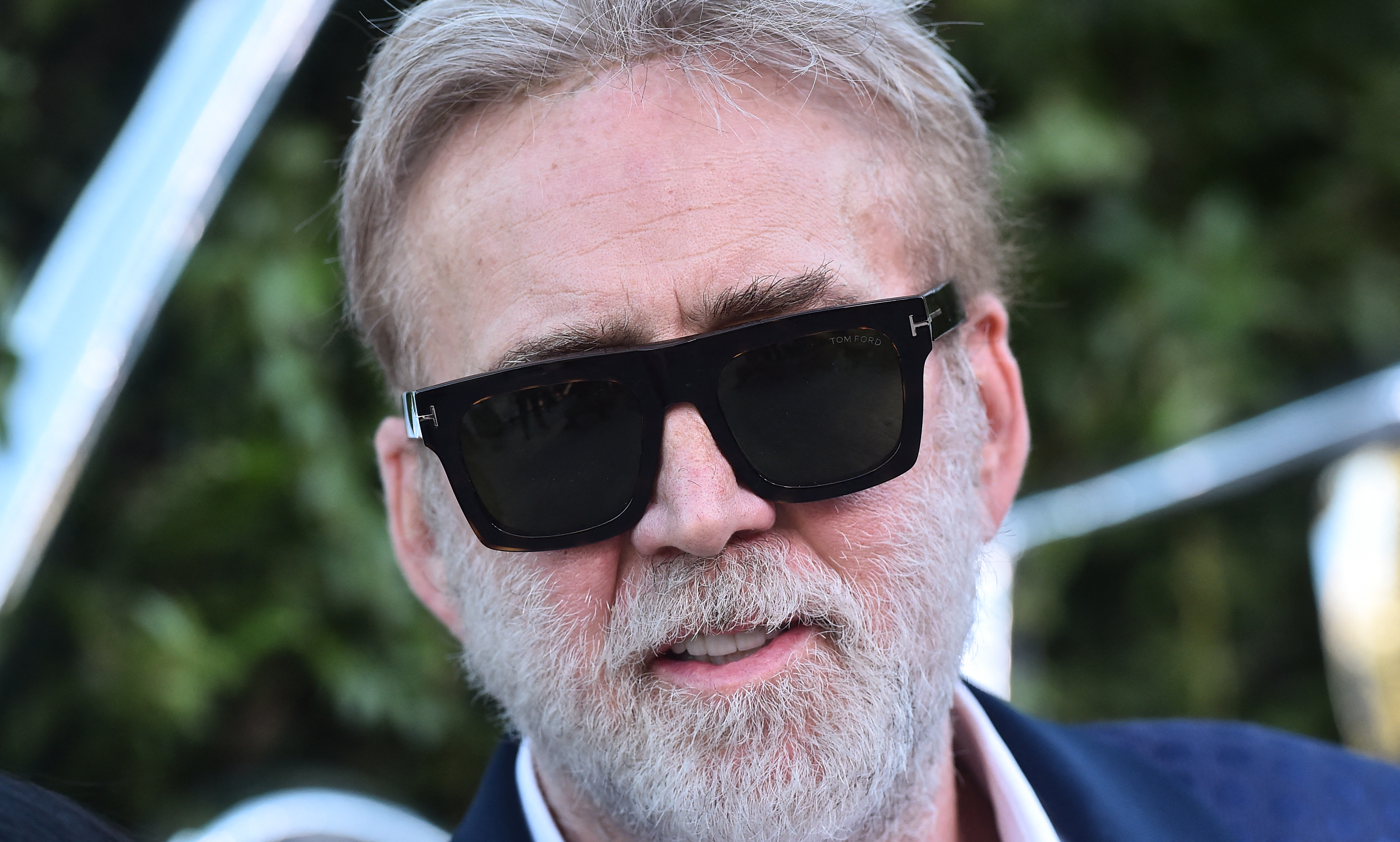 Close-up of a middle-aged man with short gray-blond hair and a gray beard wearing dark Tom Ford sunglasses and a navy blazer.