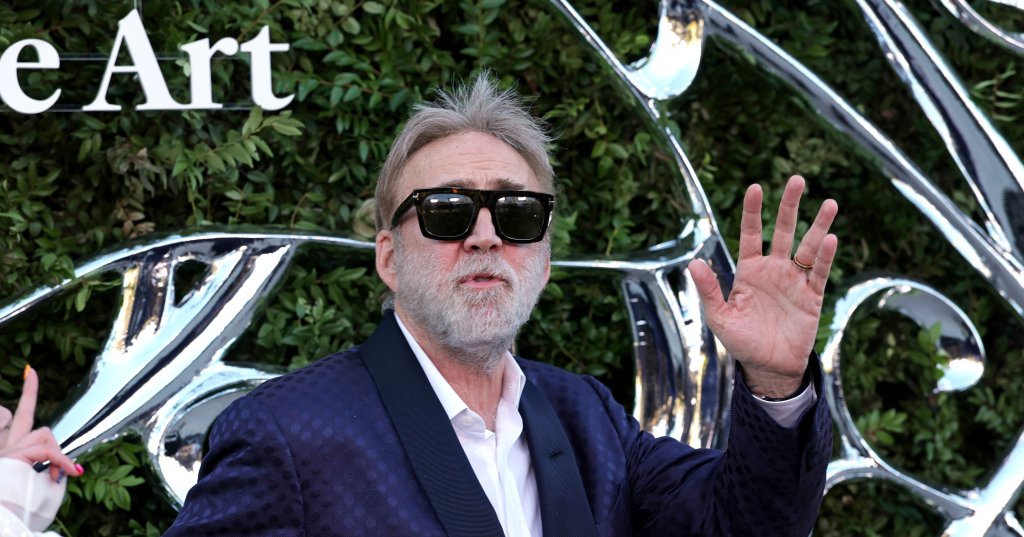 Nicolas Cage in a navy blazer and dark sunglasses raising one hand in a wave at a red carpet event.