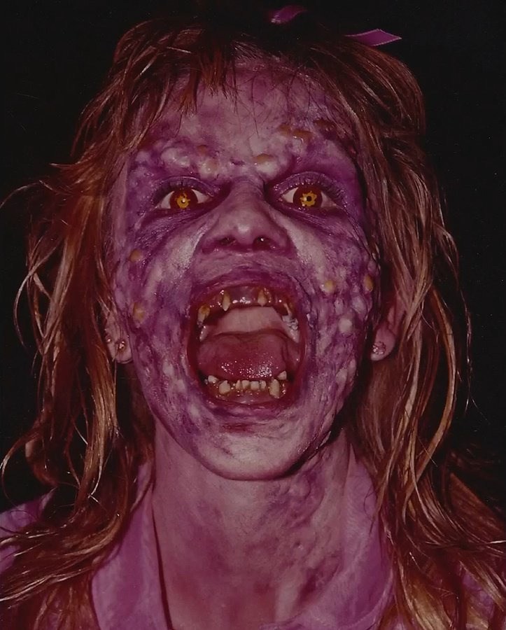 Close-up of a demon character with purple textured skin, glowing yellow eyes, open mouth with jagged teeth, and red hair with a bow.