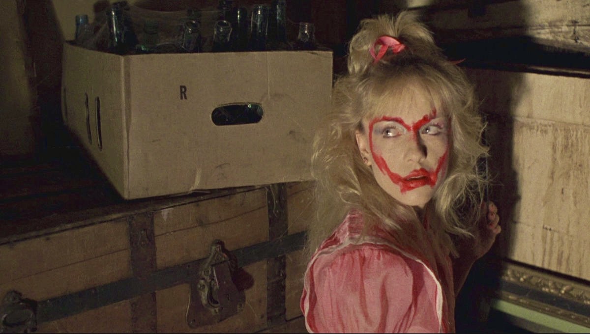 A blonde woman with red paint smeared across her face in a heart shape glances back over her shoulder in a dim, cluttered room with wooden trunks and bottles.