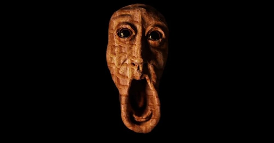 Hand-carved wooden face sculpture with wide glass eyes and a long, gaping open mouth against a black background.