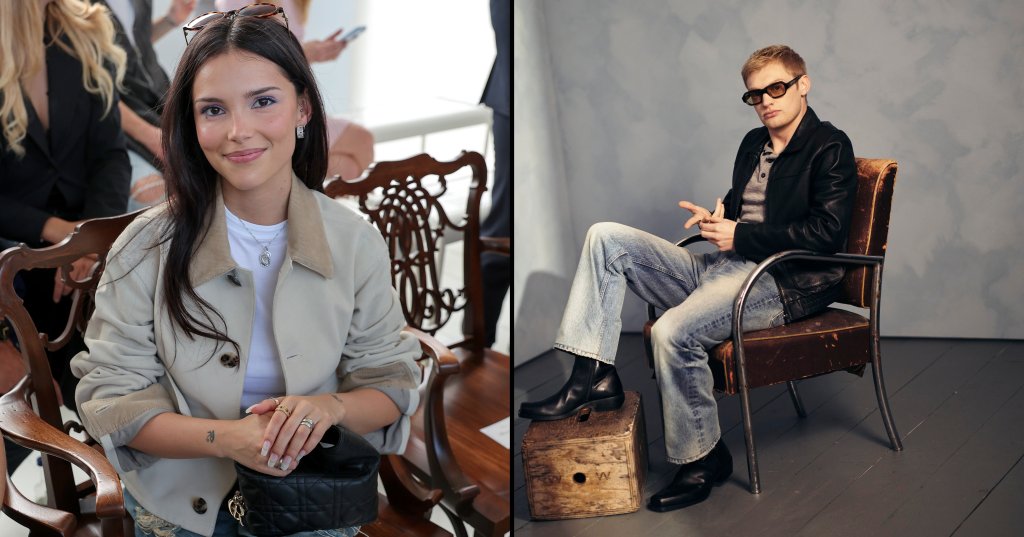 Dark-haired woman in a beige jacket and ripped jeans seated in a wooden chair at a fashion show, smiling at the camera.