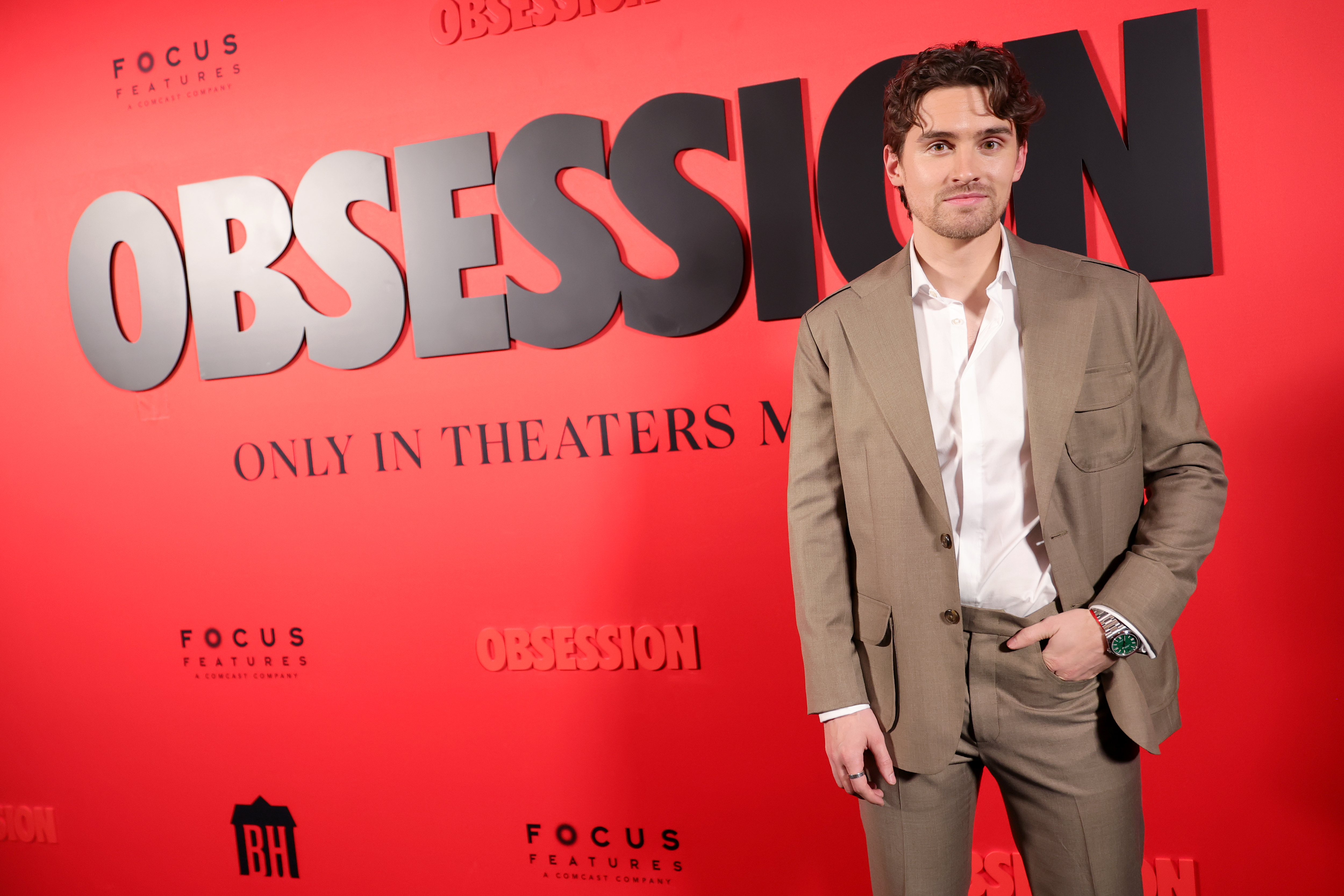 A young man in a tan suit and white shirt stands in front of an Obsession movie premiere backdrop reading 'Only in Theaters.'