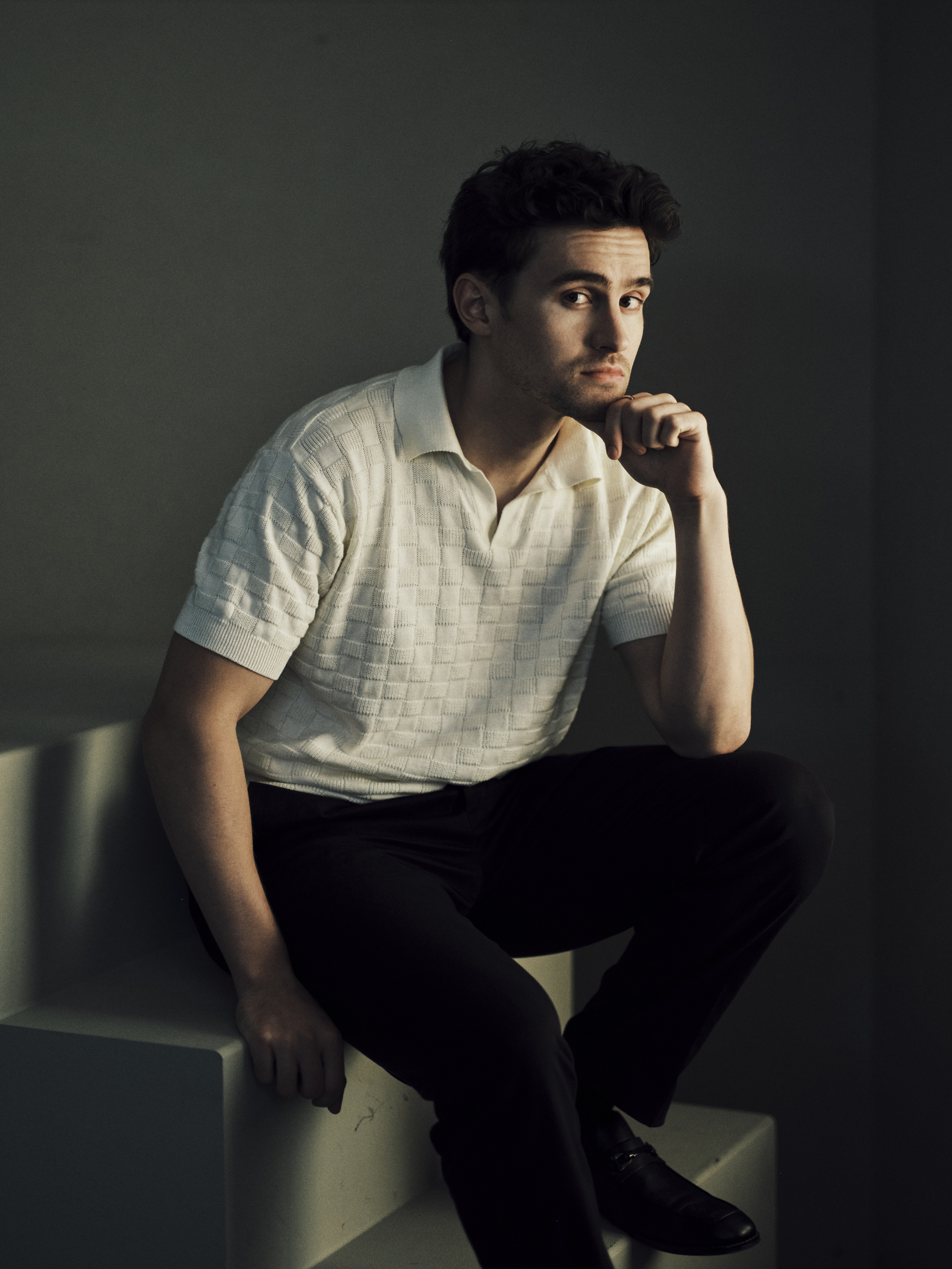 Young man with dark curly hair in a cream textured polo shirt and black trousers, seated on a white cube with his chin resting on his hand, against a dark gray background.