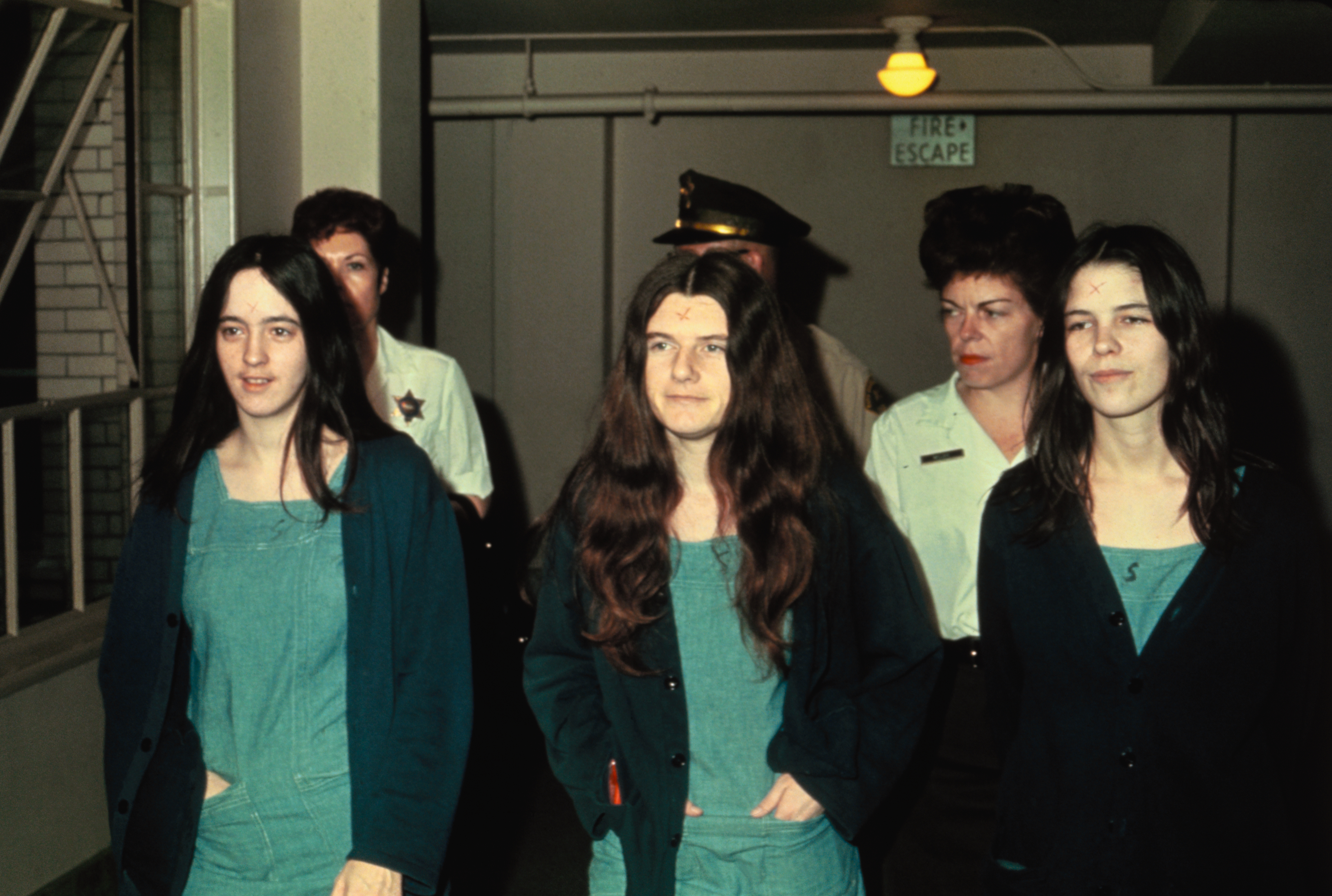 Color photo of three young dark-haired women in teal jumpsuits with X marks carved on their foreheads, escorted by law enforcement officers through a hallway with a fire escape sign overhead.