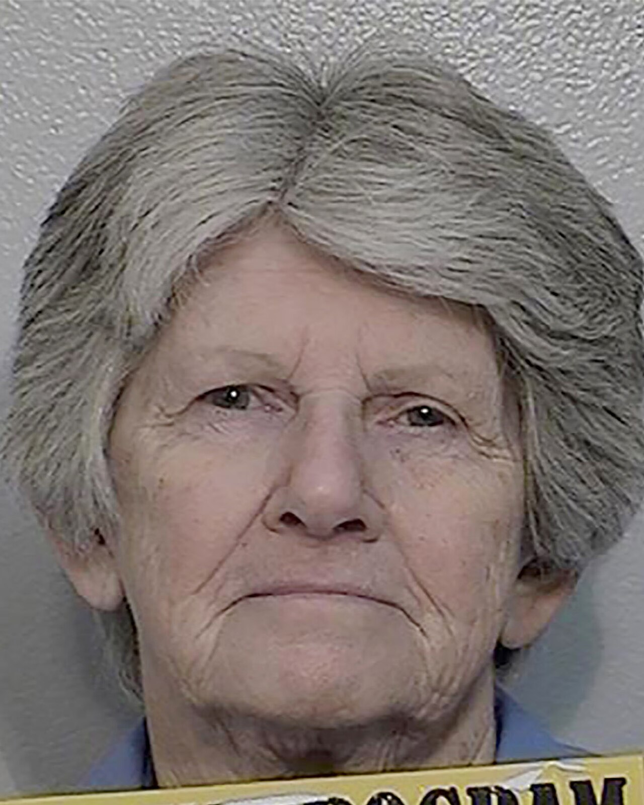 Mugshot of an elderly woman with short gray hair against a light gray wall, with a prison program placard at the bottom of the frame.
