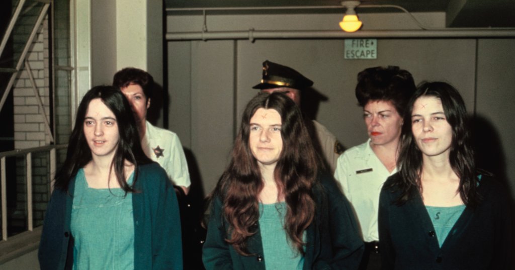 Three young women in teal denim jumpsuits with X marks on their foreheads walk escorted by uniformed deputies through a hallway.