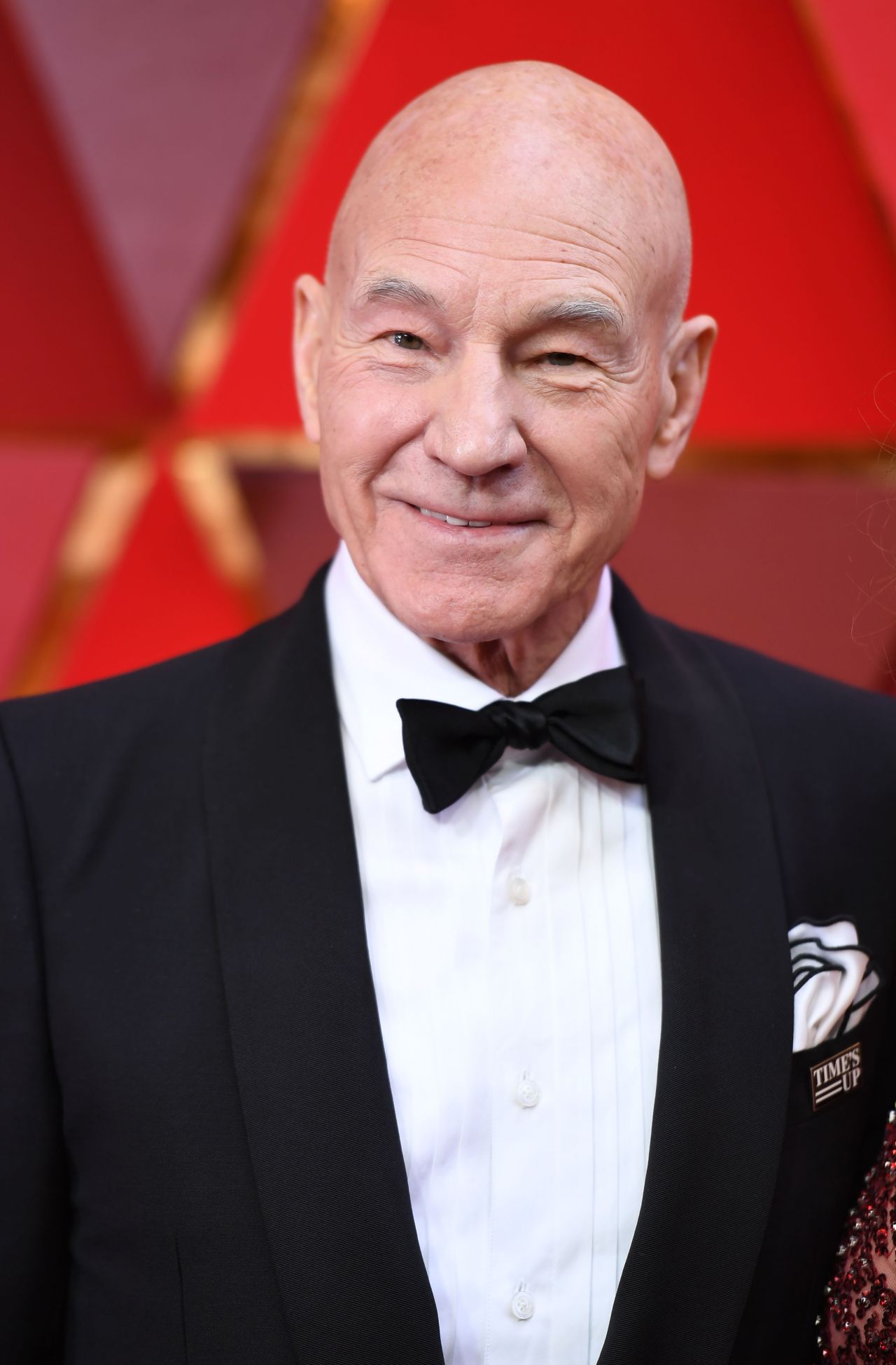 Bald man in a black tuxedo with bow tie and a Time's Up pin, smiling on a red carpet with a red decorative backdrop.