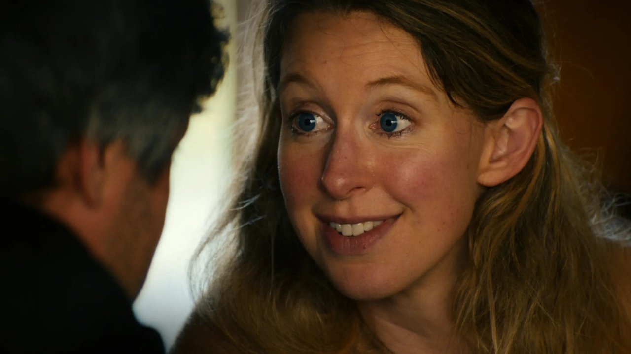 Close-up of a smiling woman with blue eyes and long blonde-brown hair looking at someone out of frame.